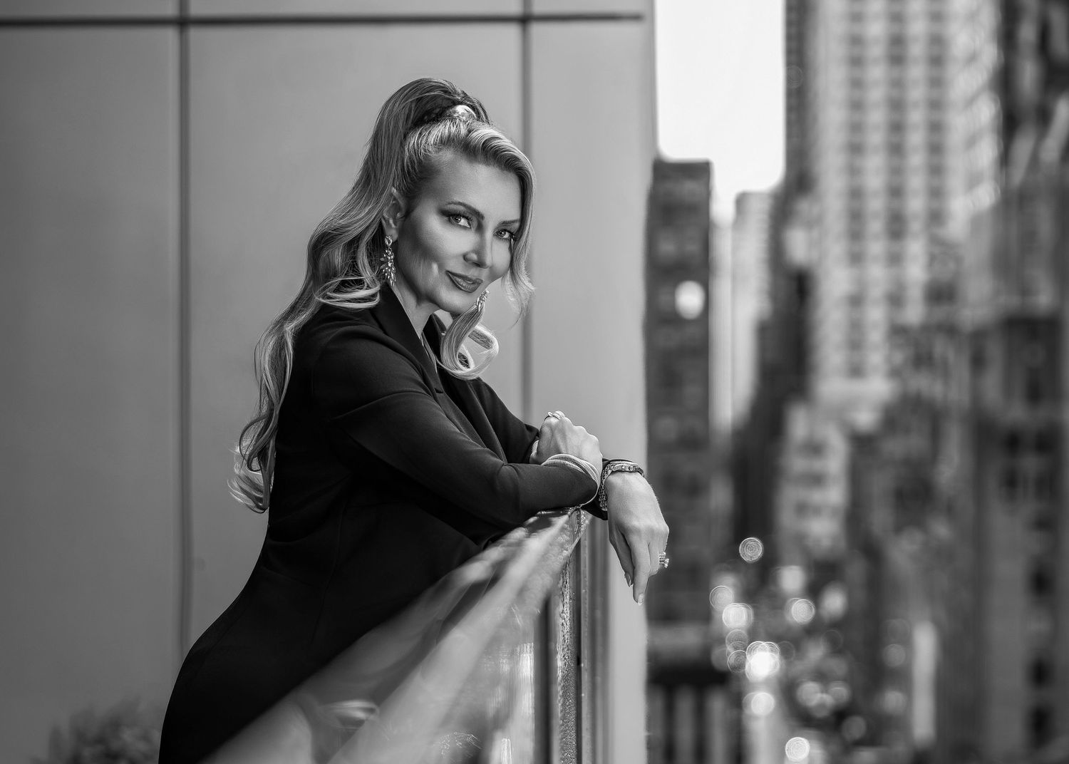 Black and white photo of a smiling blonde woman leaning on a railing with a city skyline backdrop.