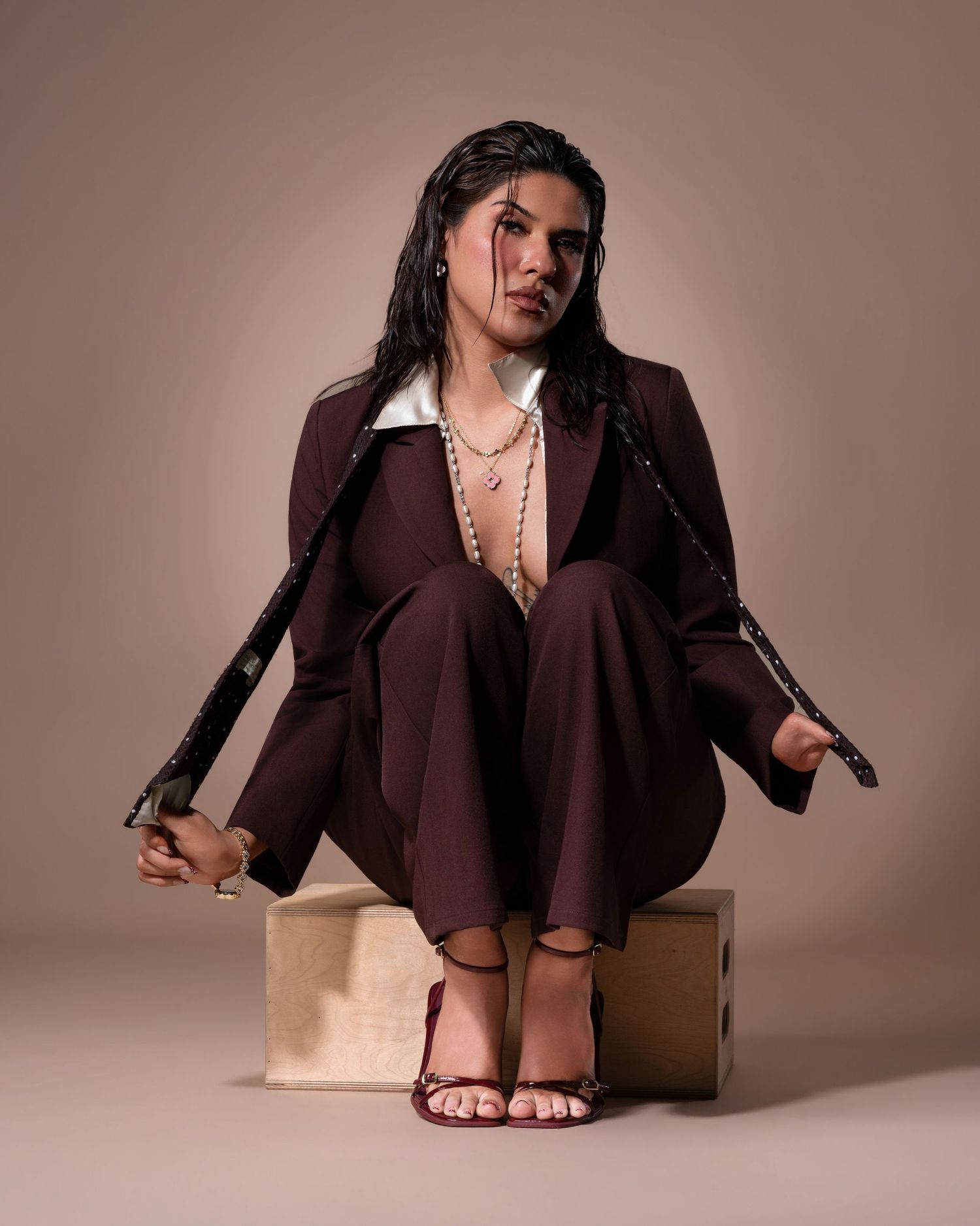 Person in brown suit seated on wooden box, wearing heels and layered necklaces, with braided hair accents.
