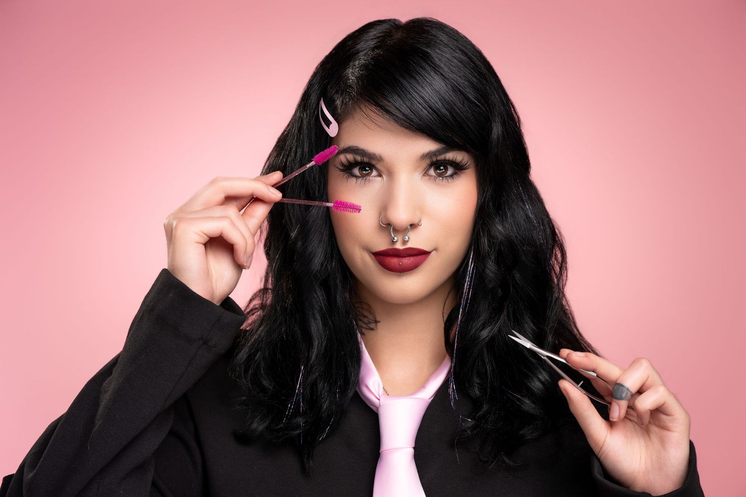 Young woman with black hair and nose ring holding pink tweezers and scissors against a pink background.