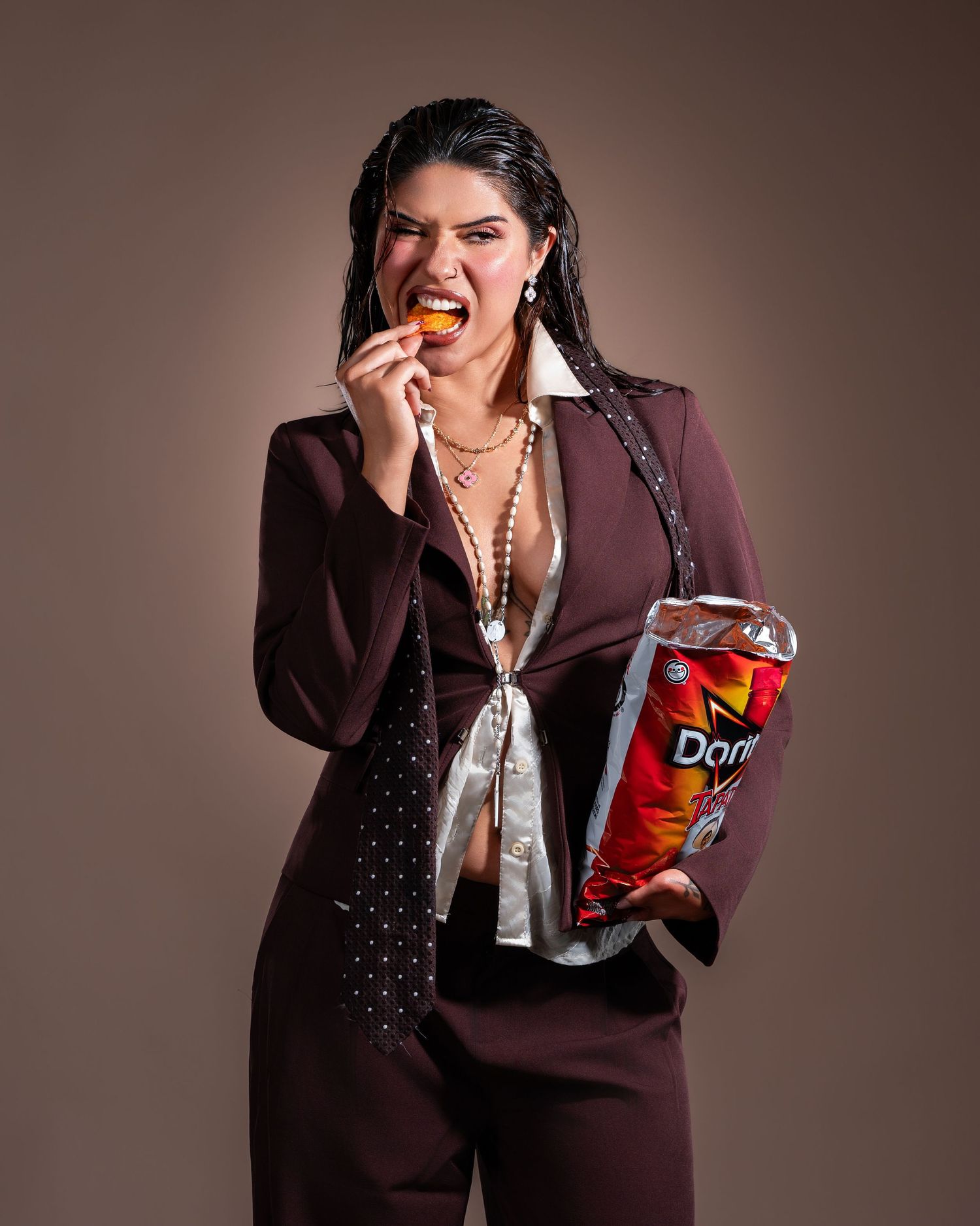 Woman in maroon suit eating Doritos chip while holding large bag, posing confidently against brown background.