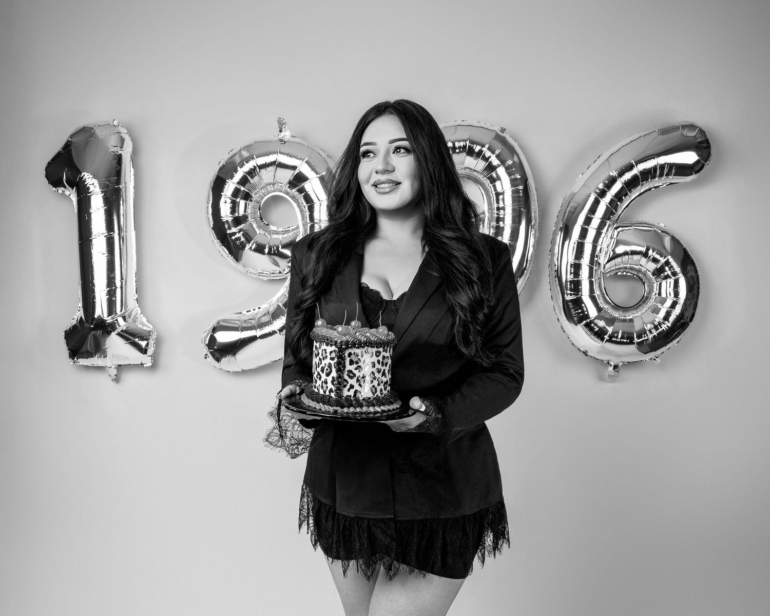 Woman holding a decorated birthday cake in front of large silver number balloons spelling 1996, in black and white.