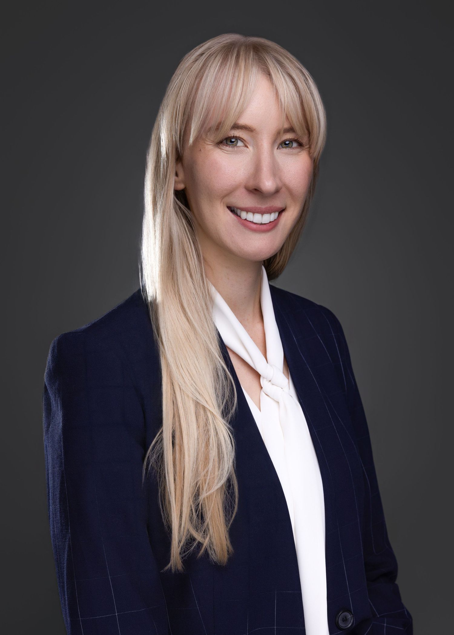 Professional headshot of a smiling blonde woman in a navy blazer and white blouse against a dark gray background.