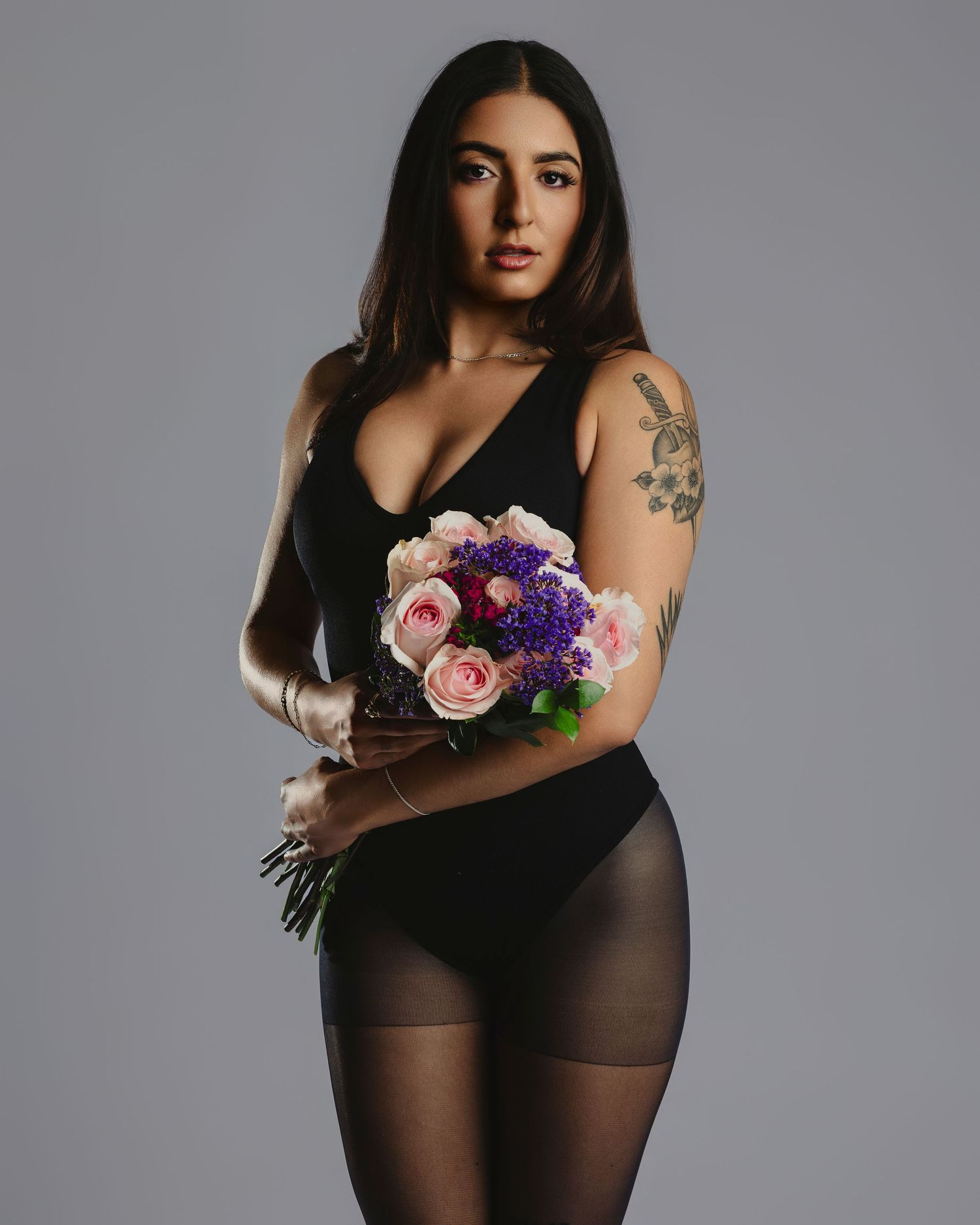 Woman in black bodysuit and tights holds pink and purple flower bouquet, showcasing arm tattoo in studio portrait.