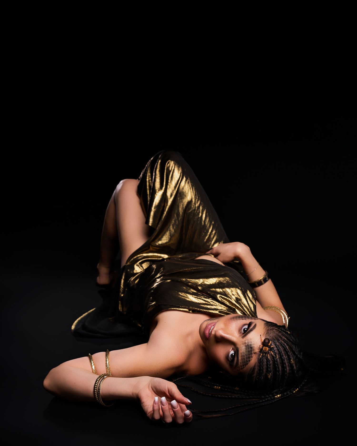 Woman in gold metallic dress lying on dark floor, wearing gold jewelry with braided hair.