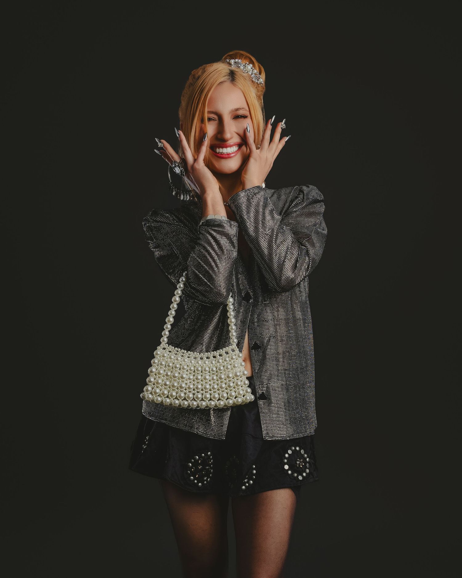 Smiling blonde woman poses in gray blazer, black studded skirt, pearl handbag, and tiara against dark background.