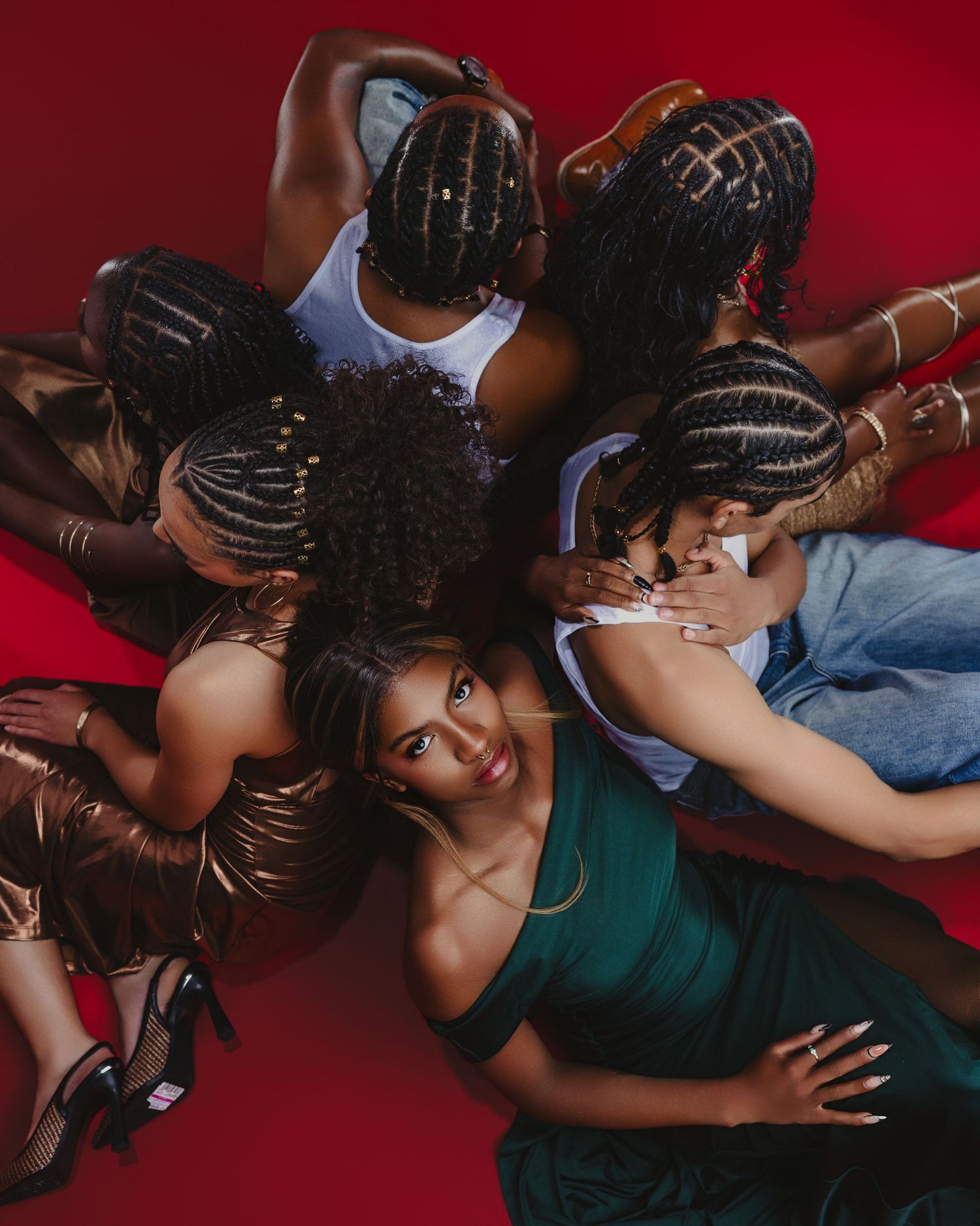 Group of women with braided hairstyles posing together in a circle on a red background, wearing colorful outfits.