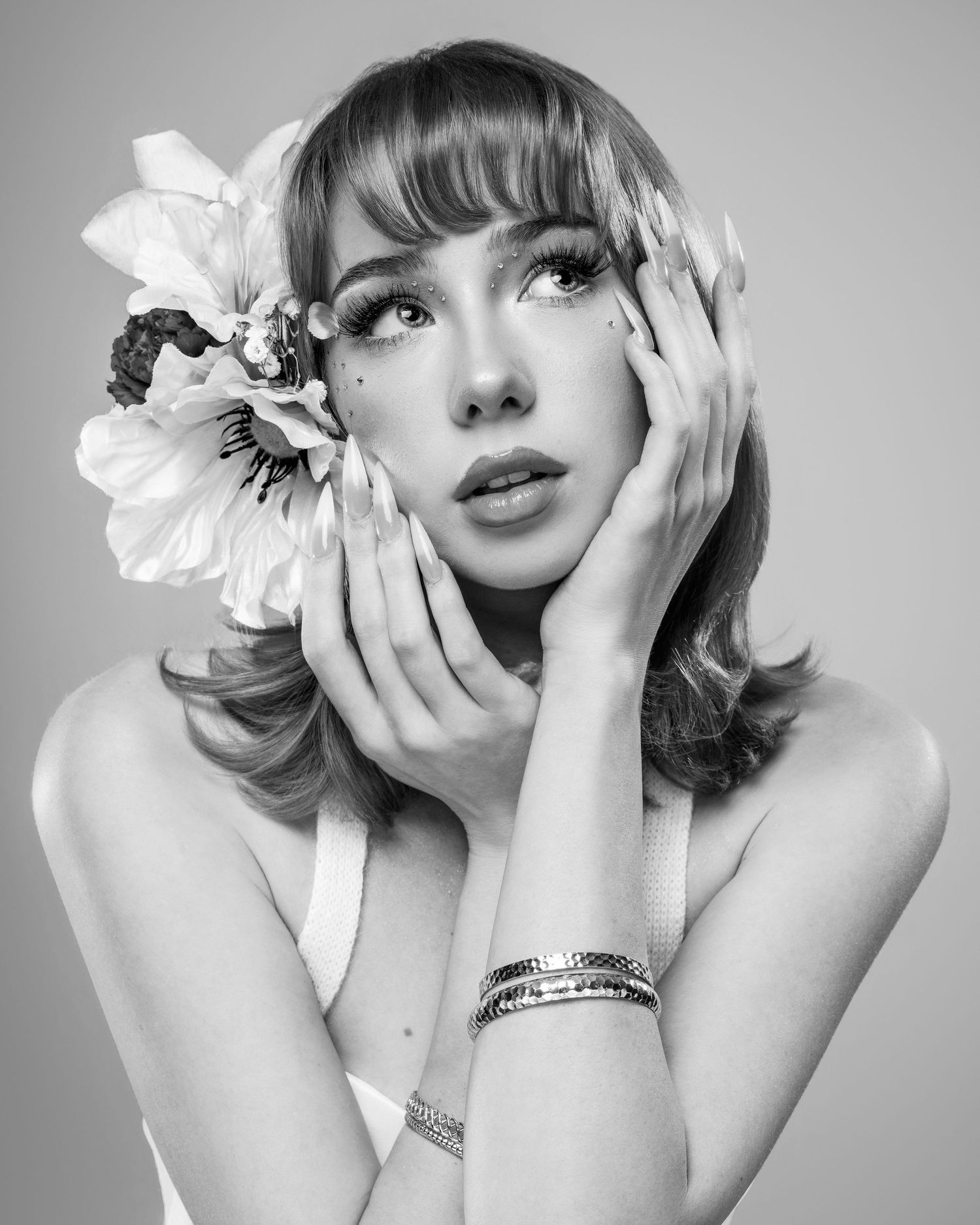 Black and white portrait of a woman with flower hair accessory, bracelets, and elegant pose against gray background.