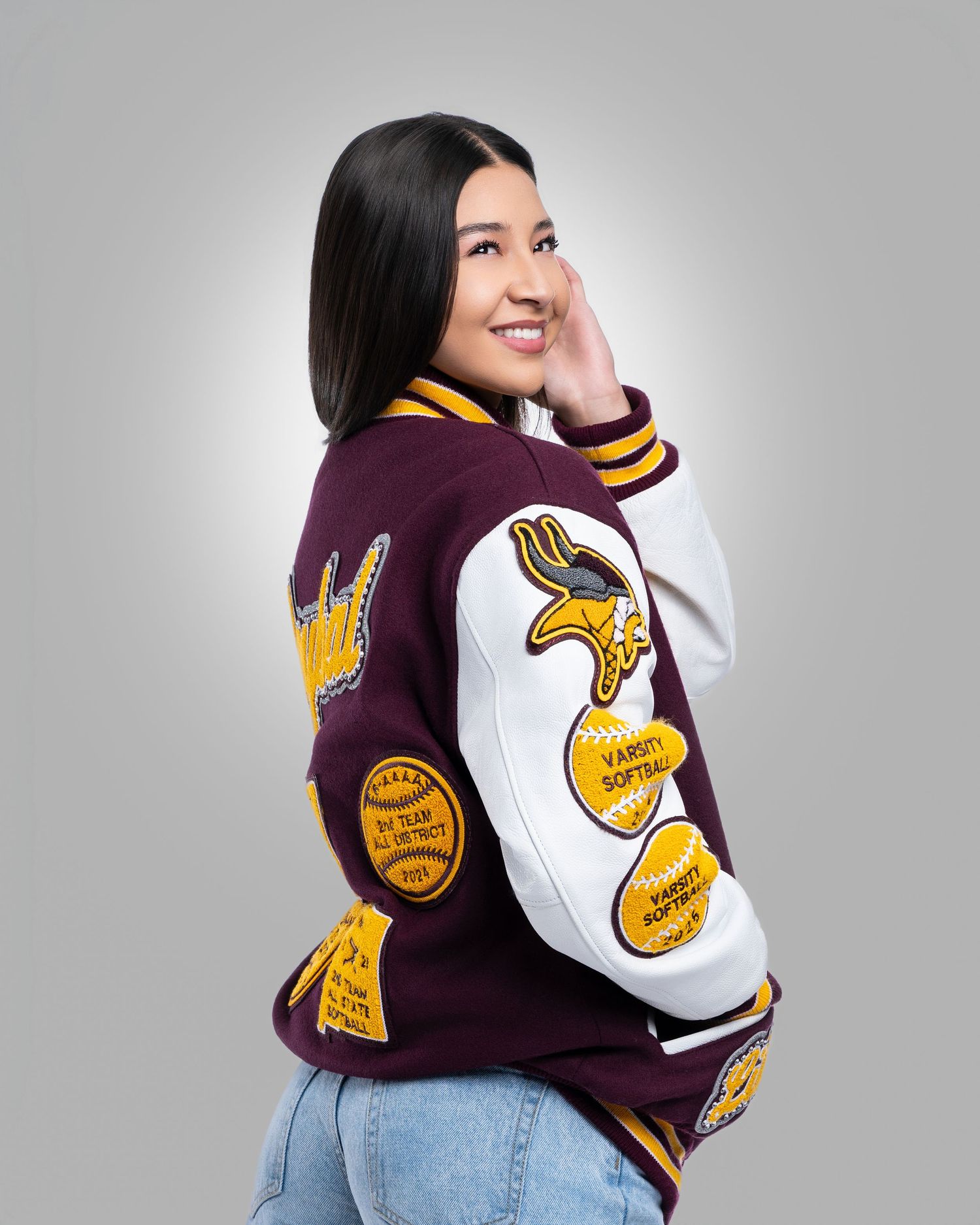 Young woman wearing a maroon and white varsity softball letterman jacket posing in a studio.