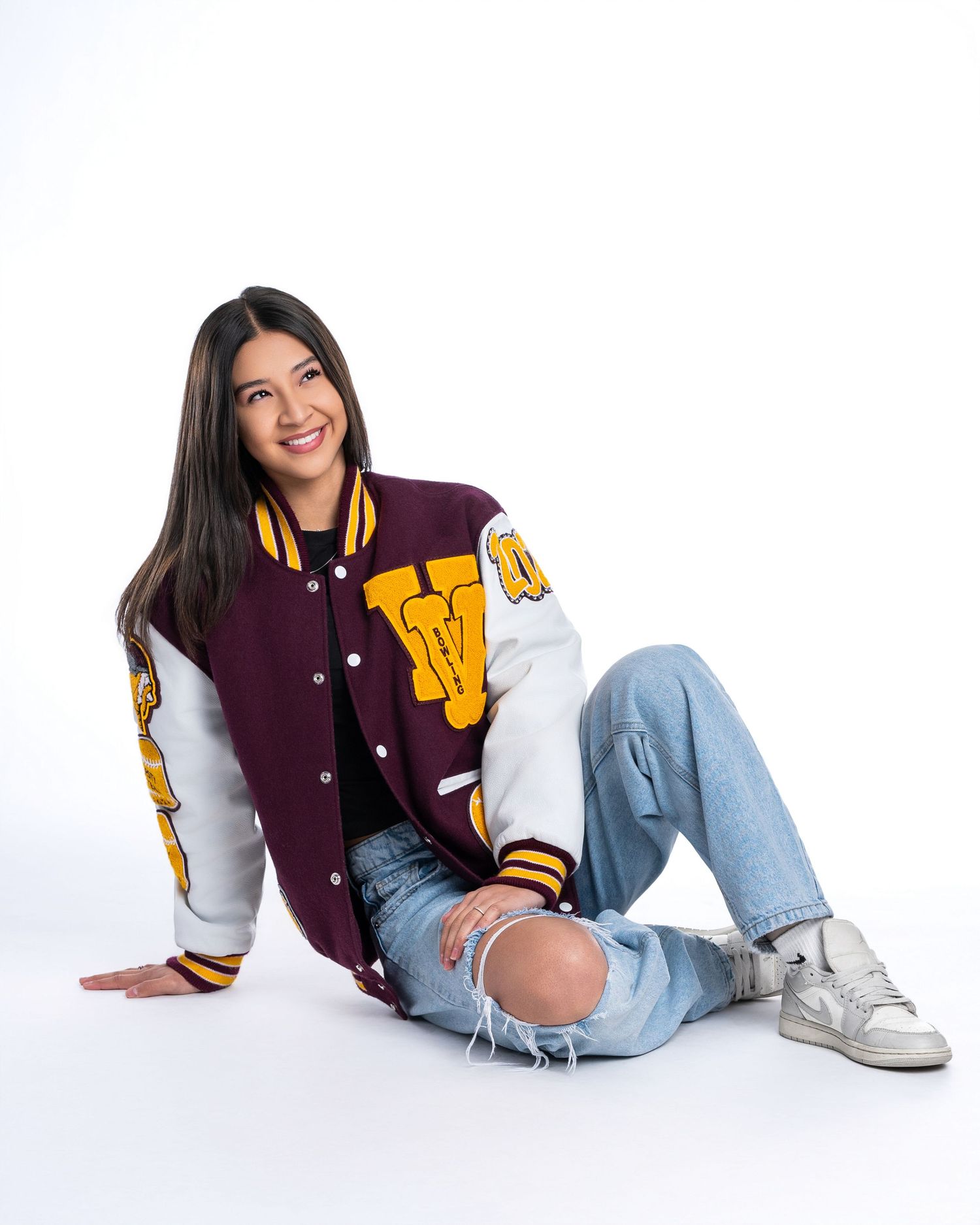 Young woman in maroon varsity jacket and ripped jeans sits on white studio floor smiling.