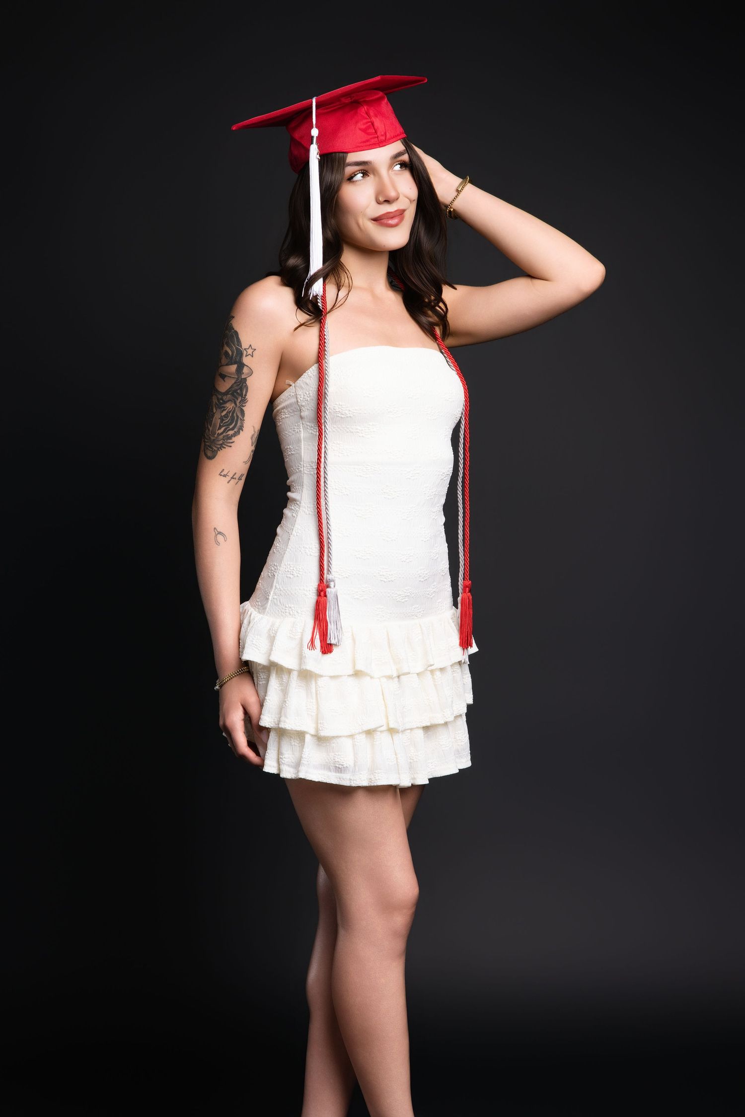 Young woman in white dress and red graduation cap posing in studio with tattooed arm visible.