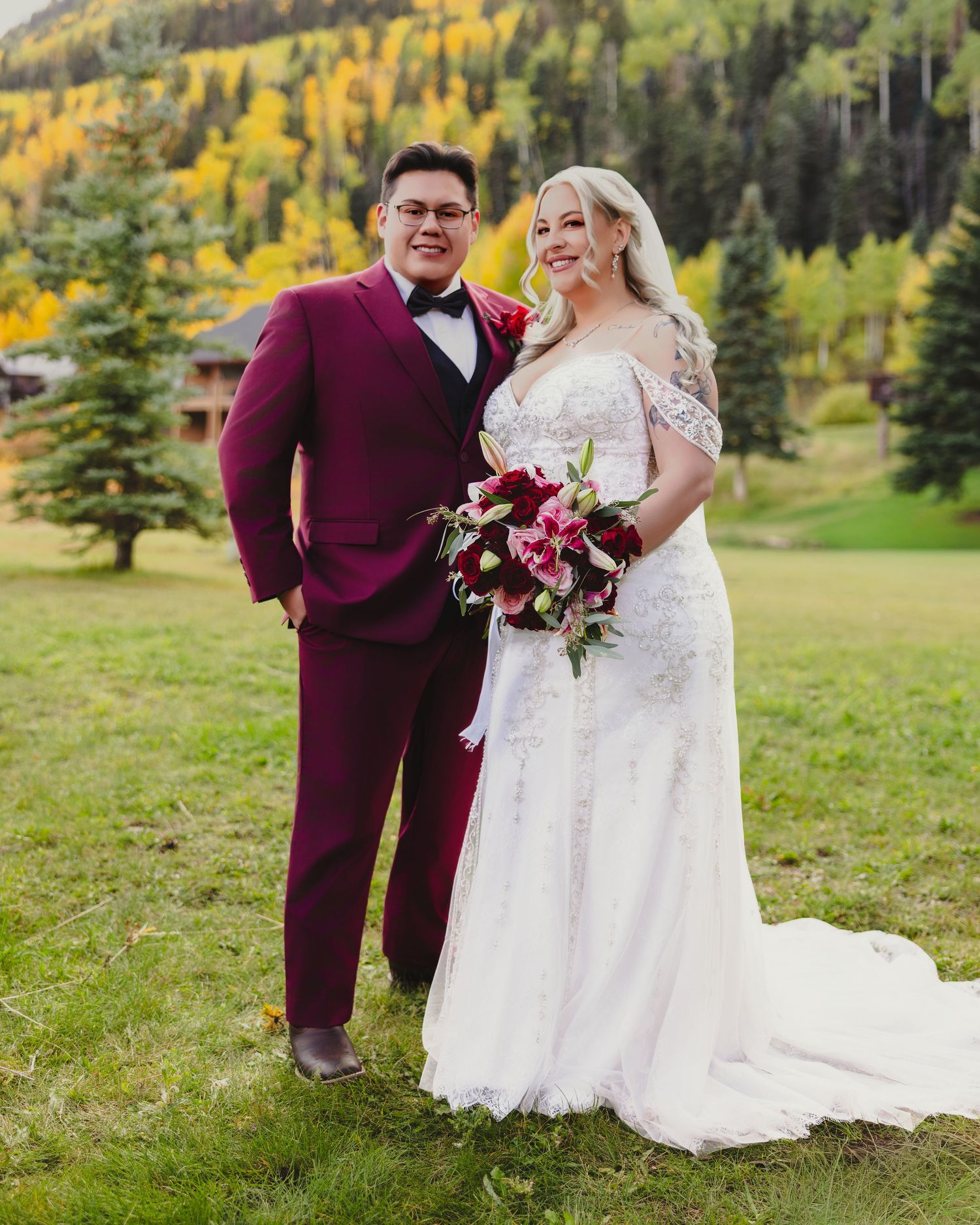 Smiling bride and groom posing in burgundy suit and white lace gown at fall mountain wedding.