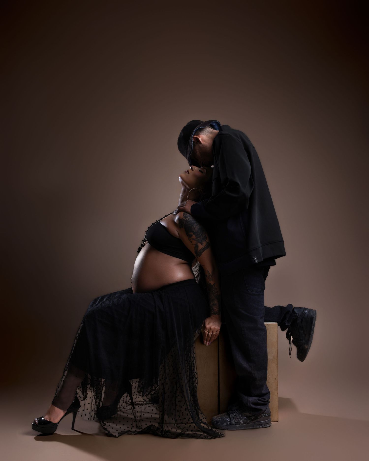 Couple during maternity photoshoot, man kissing pregnant woman's belly, both dressed in black.