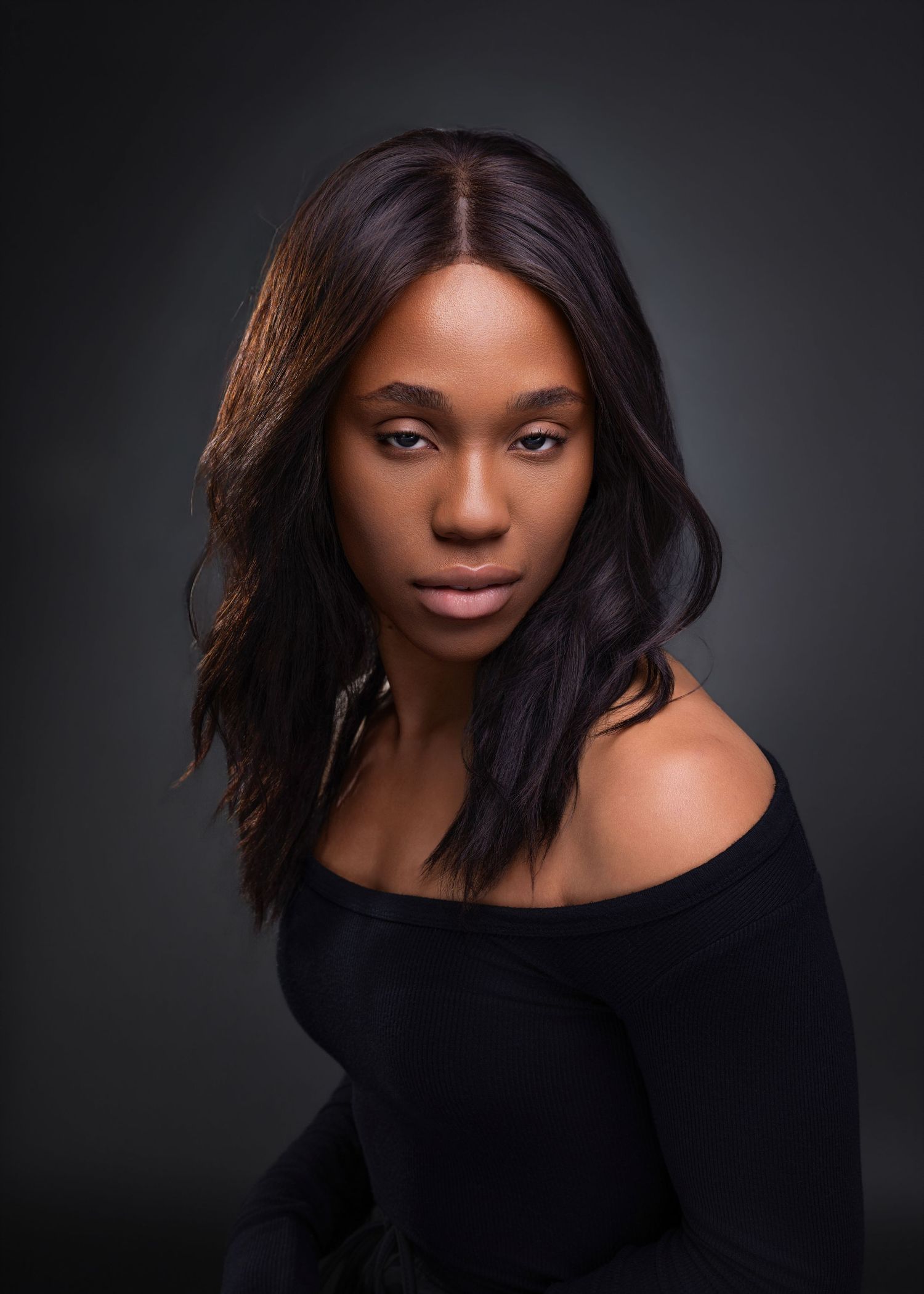 A young Black woman in an off-shoulder black top with long wavy hair poses confidently against a dark studio background.