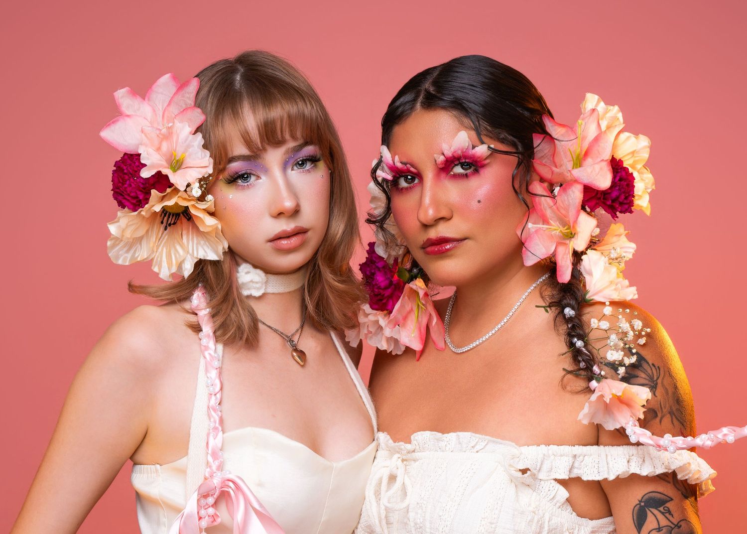 Two women with floral hair accessories and pink makeup pose together against a pink background in white dresses.