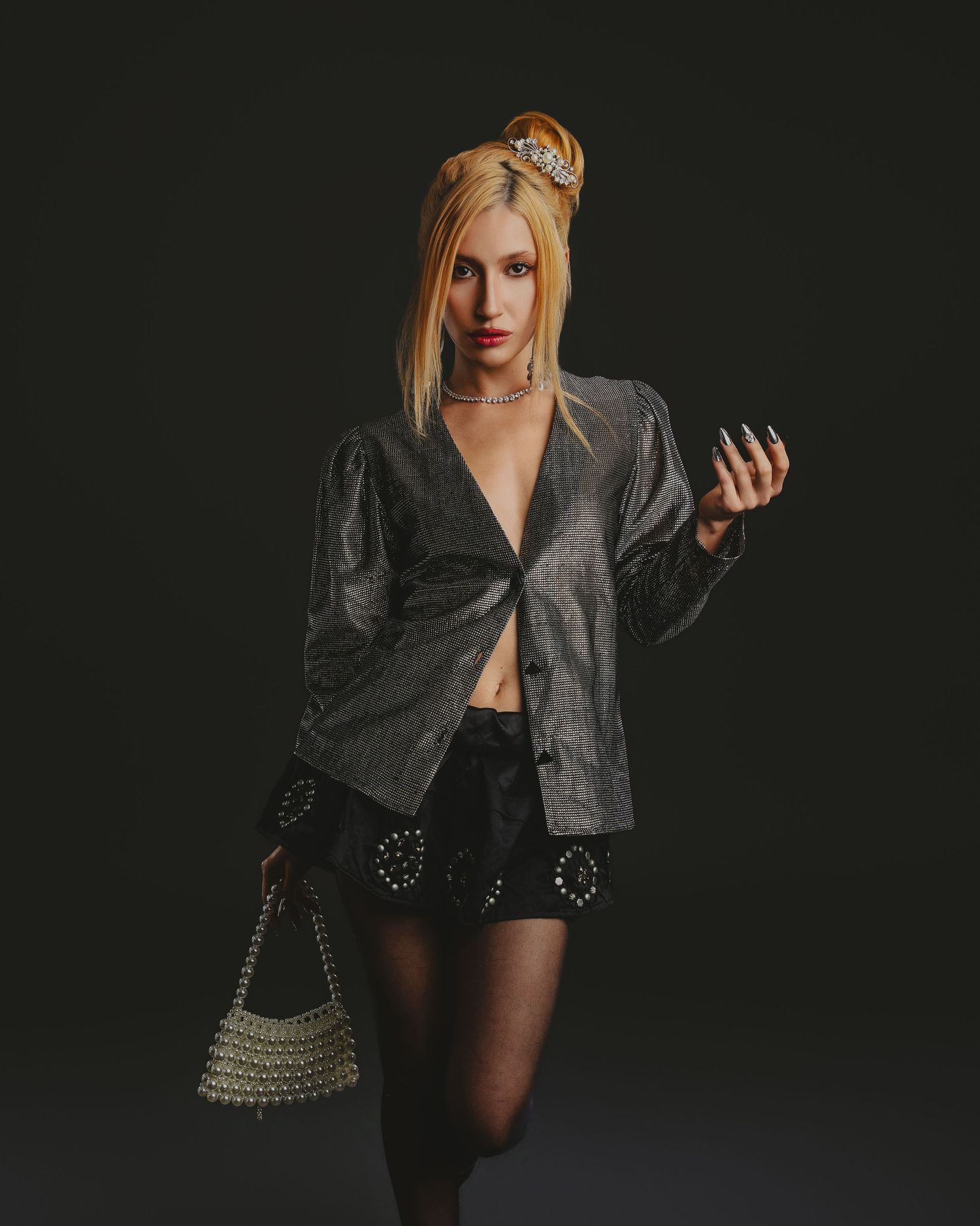 Blonde woman in silver metallic blazer, black mini skirt, sheer tights, pearl bag, and tiara poses on dark background.