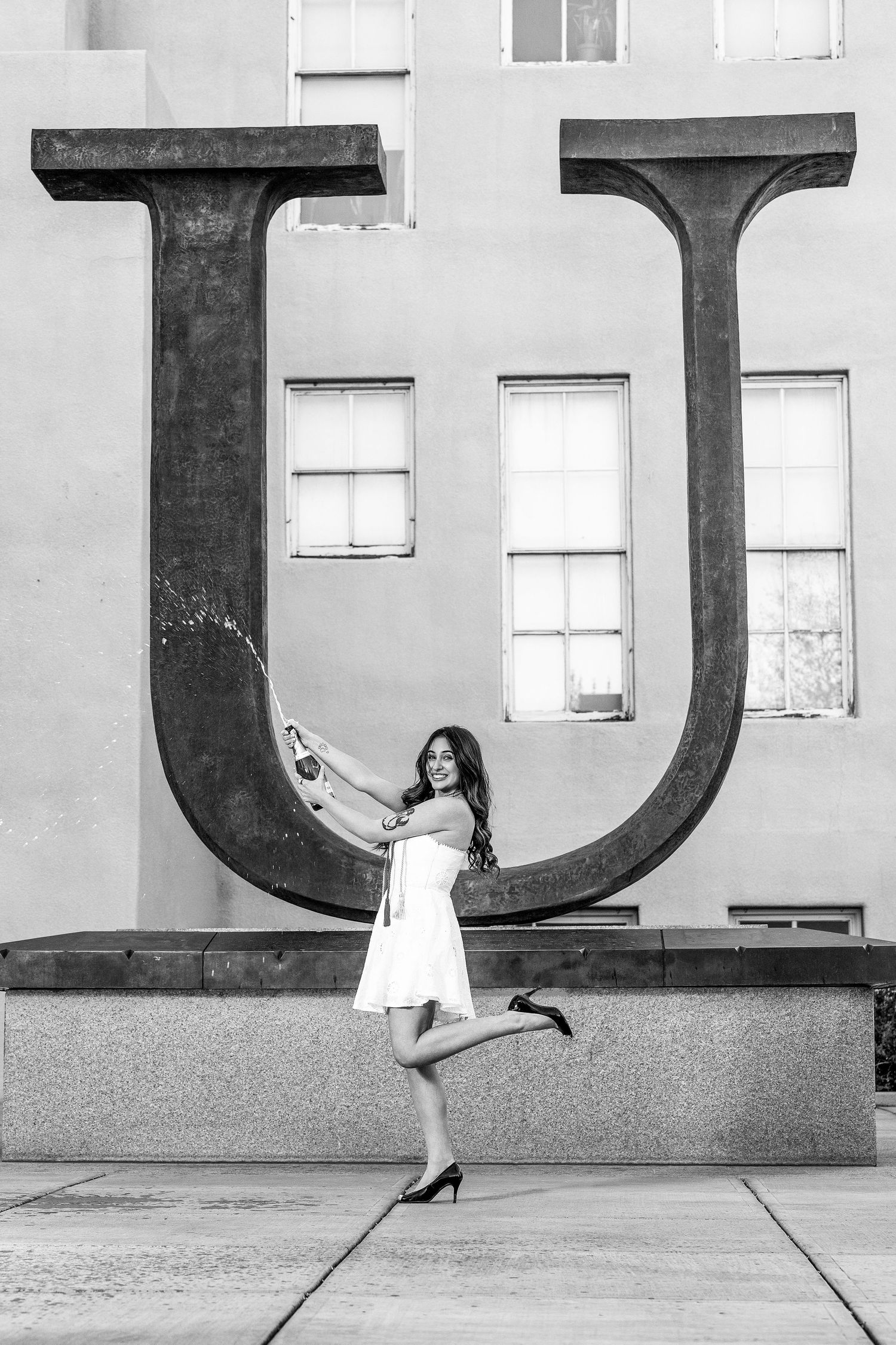 Young woman in white dress pops champagne beside large metal U sculpture in black and white.