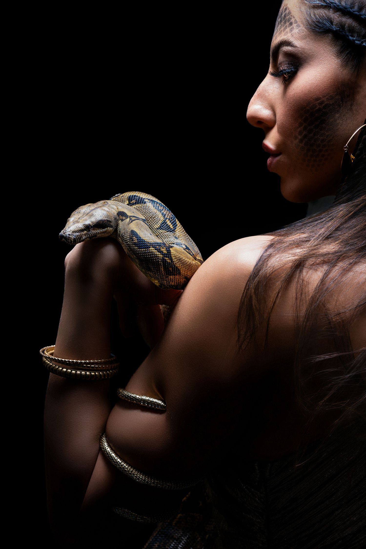 A woman with scale makeup holds a python snake against a dark background, wearing gold bracelets and jewelry.