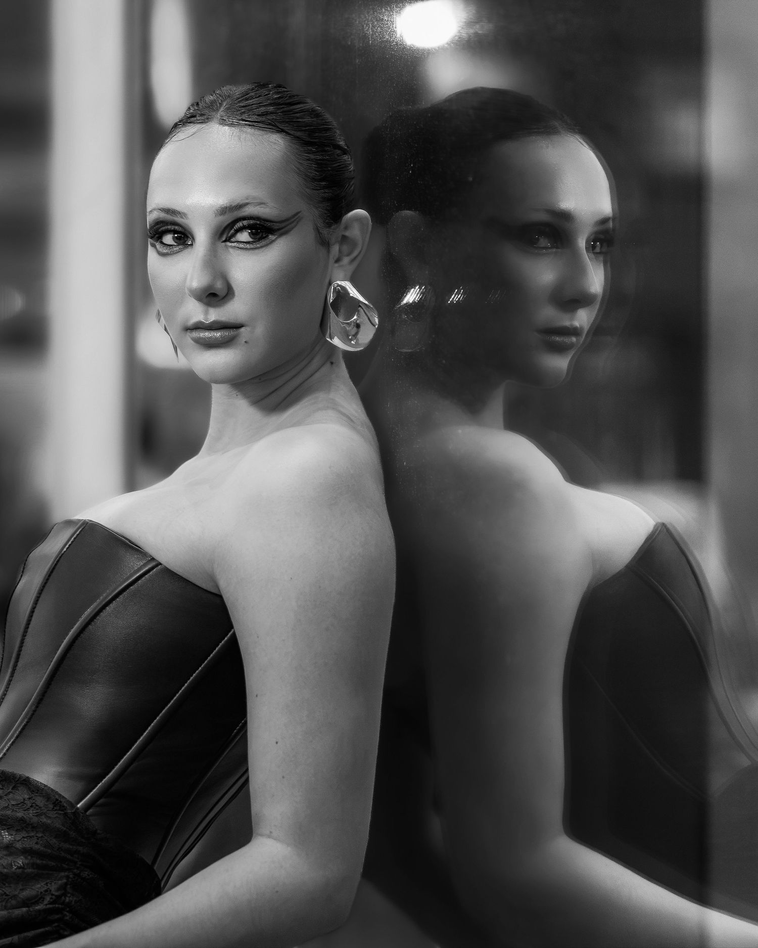 Black and white portrait of a woman in strapless leather top with bold earring, reflected in glass mirror.