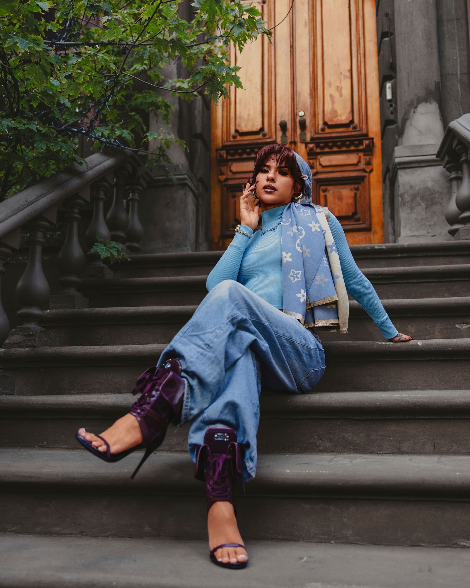 Woman in blue jeans and top with purple heeled boots sits on stone steps near ornate wooden door.