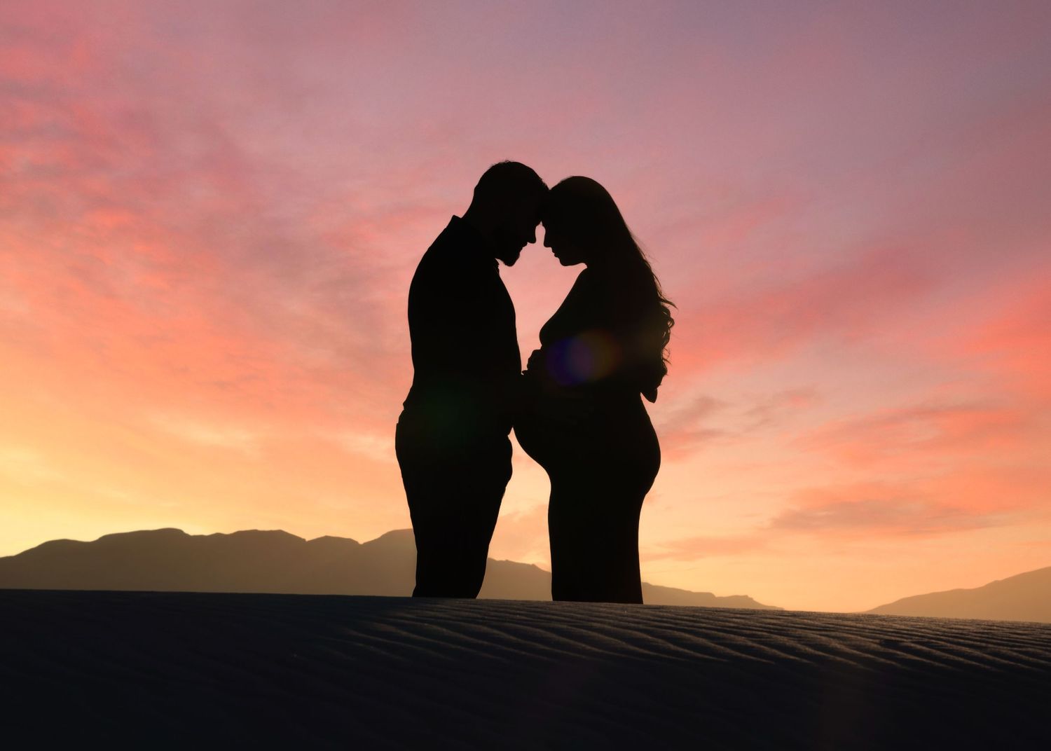 Romantic silhouette of pregnant couple touching foreheads against a vibrant pink and orange desert sunset sky.