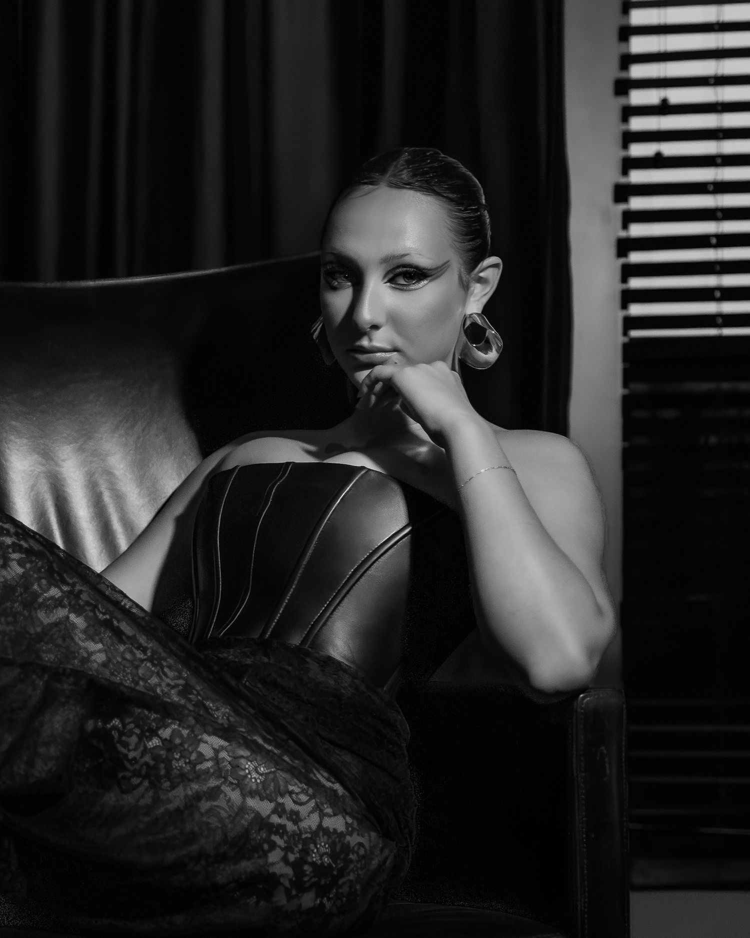 Black and white portrait of a woman in a corset top seated in a leather chair, gazing at camera with dramatic lighting.