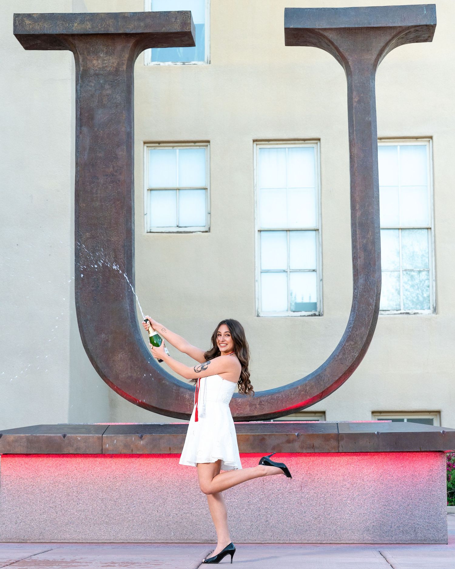 Young woman in white dress pops champagne bottle beside large metal U sculpture outdoors.