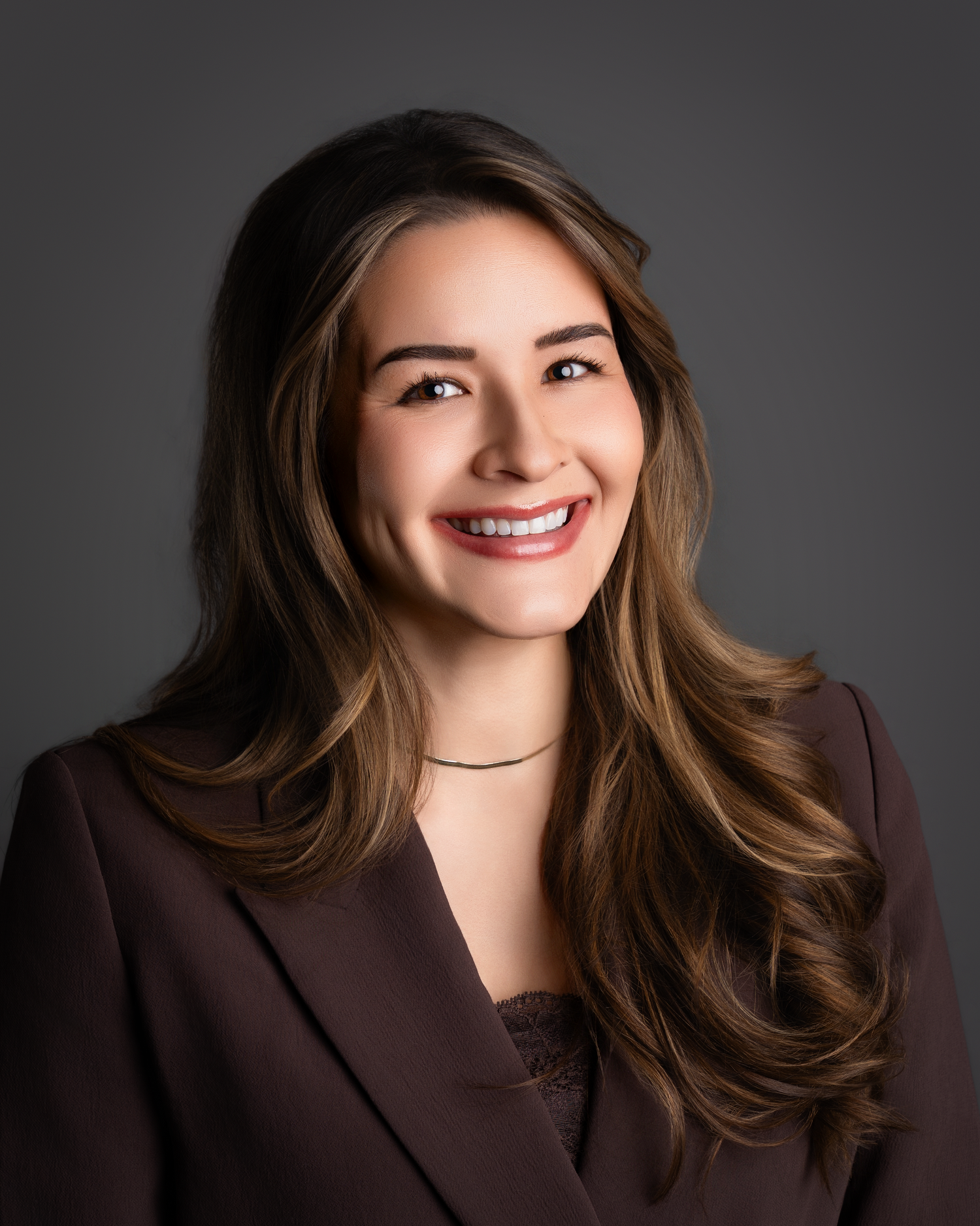 Professional headshot of a smiling young woman with long brown hair wearing a dark blazer against a gray background.
