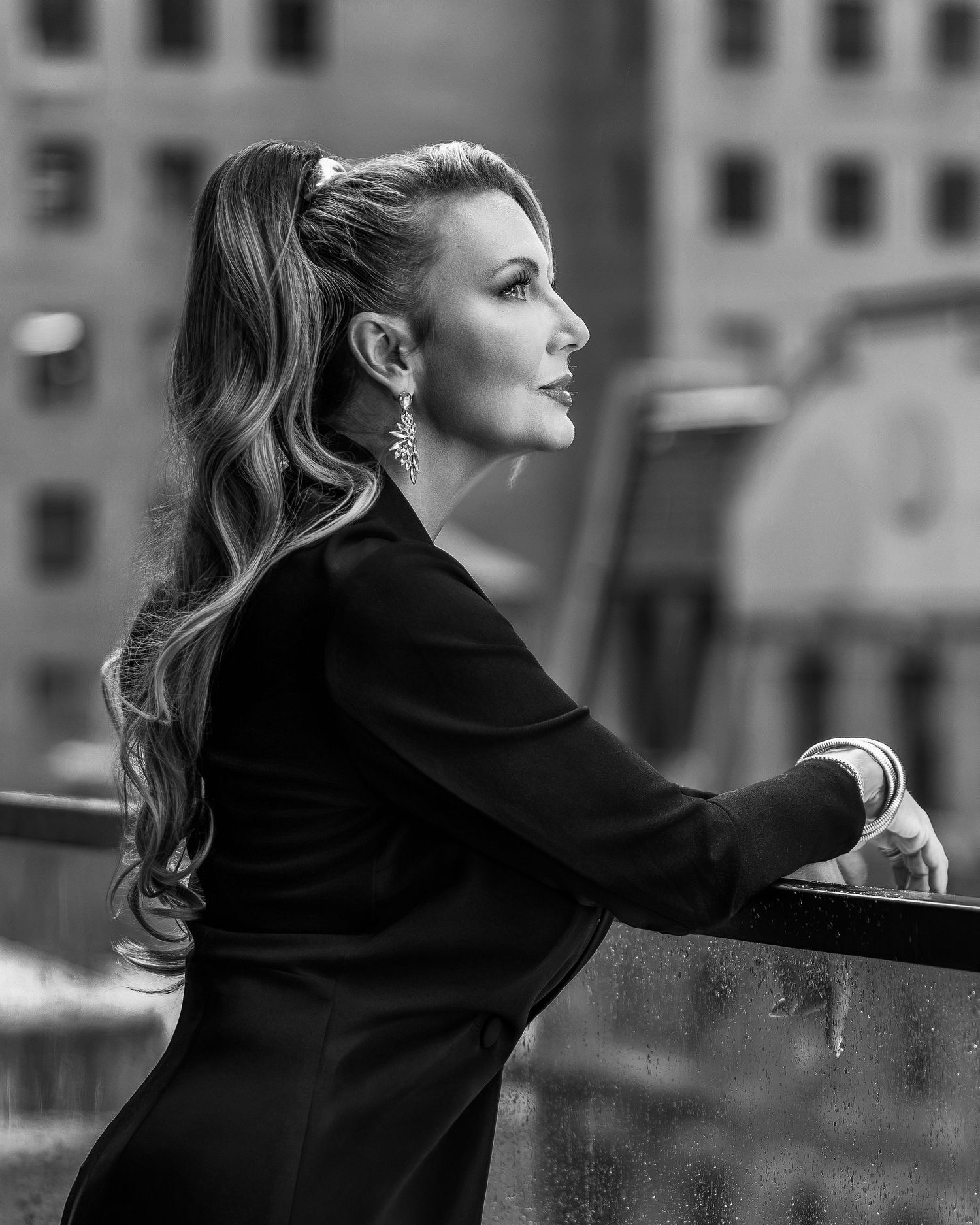 Black and white portrait of elegant woman with long wavy ponytail leaning on railing, wearing black outfit and chandelier earrings.