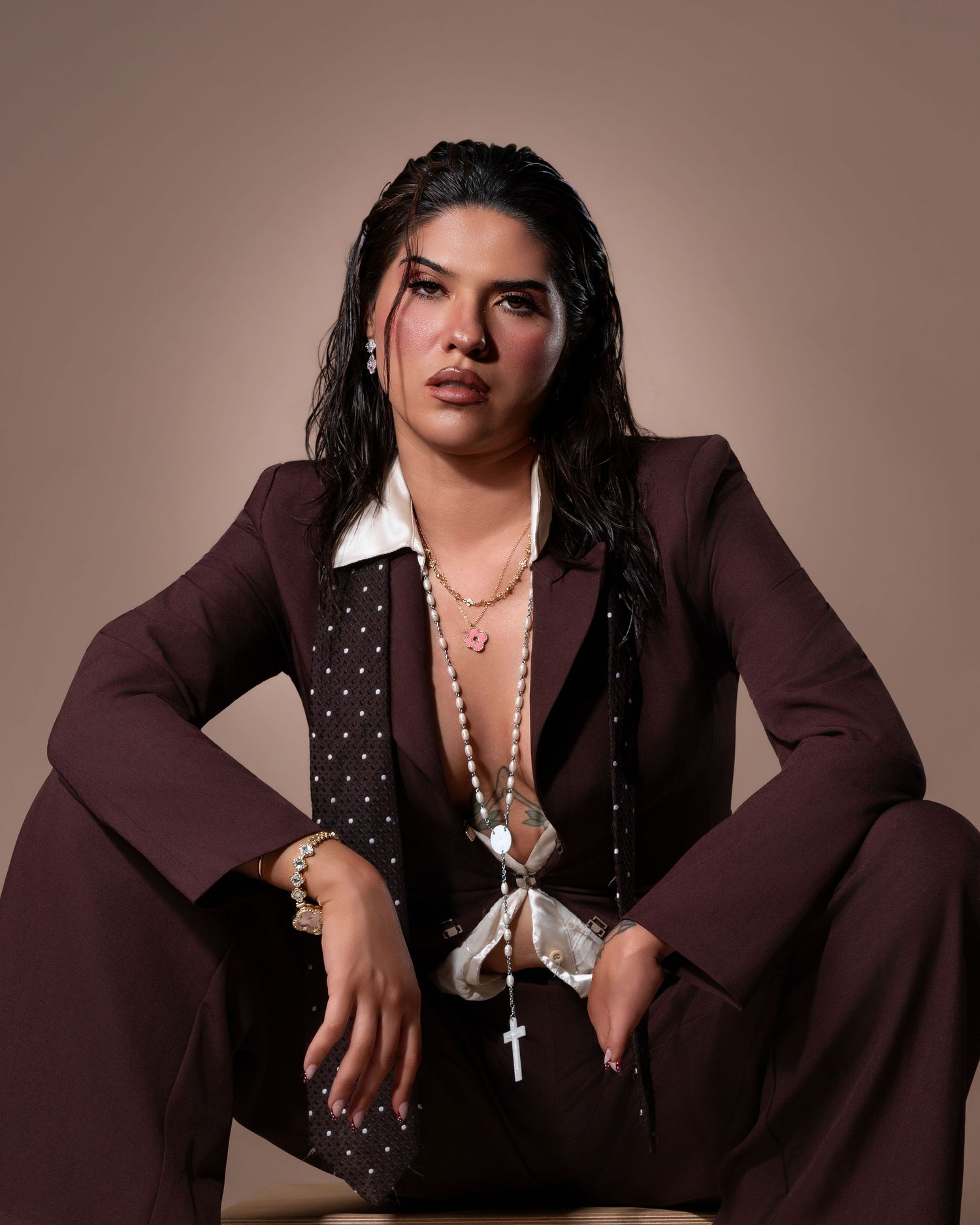 Woman in brown suit with cross necklace sits confidently against neutral background.