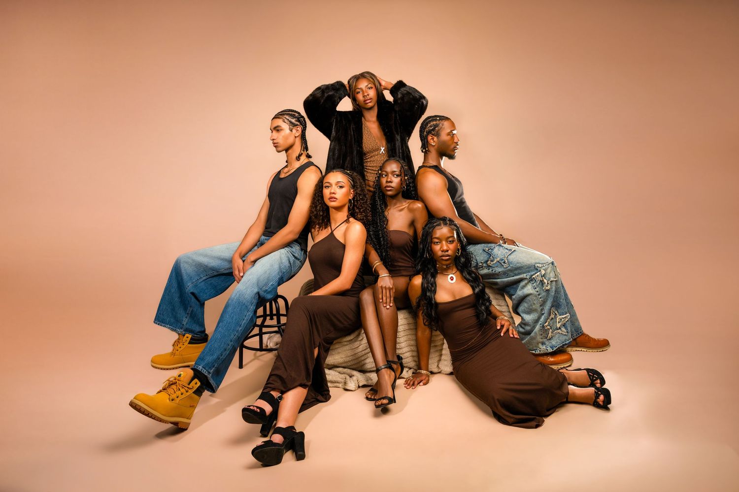 Seven Black women pose confidently together in a studio, wearing brown and denim outfits against a warm beige background.