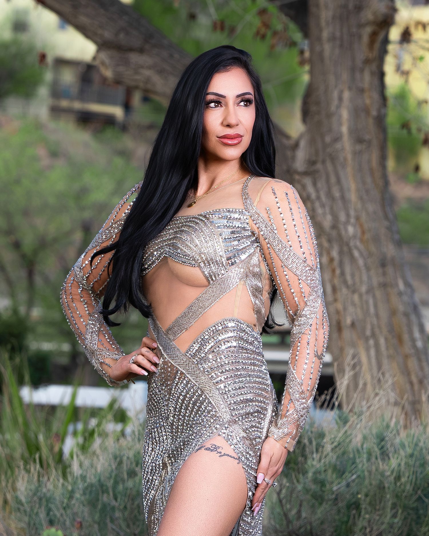 Woman in a sparkling silver rhinestone cutout dress posing outdoors near trees in a glamorous fashion photo.