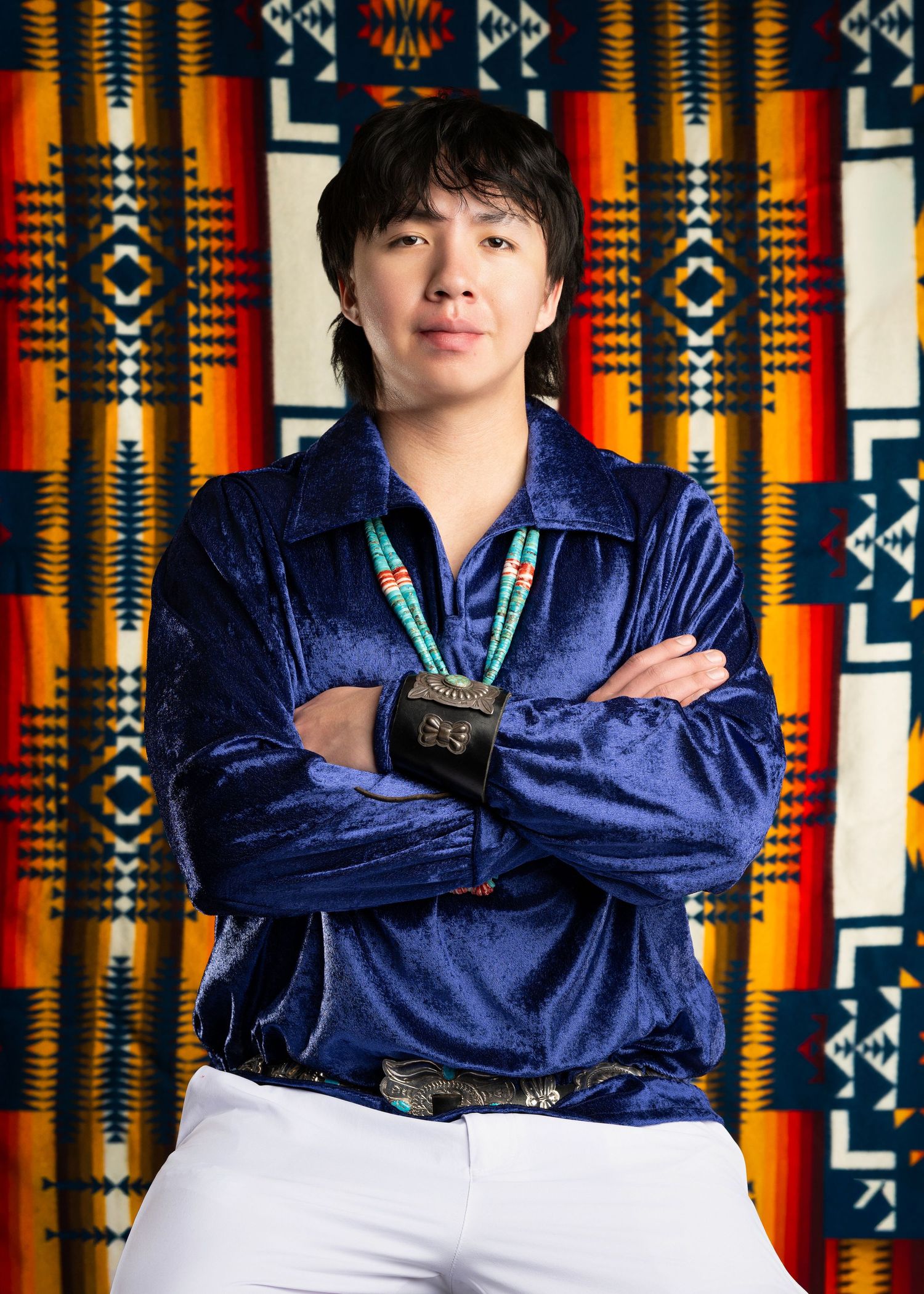Person in blue velvet shirt with arms crossed standing before a colorful Native American textile.