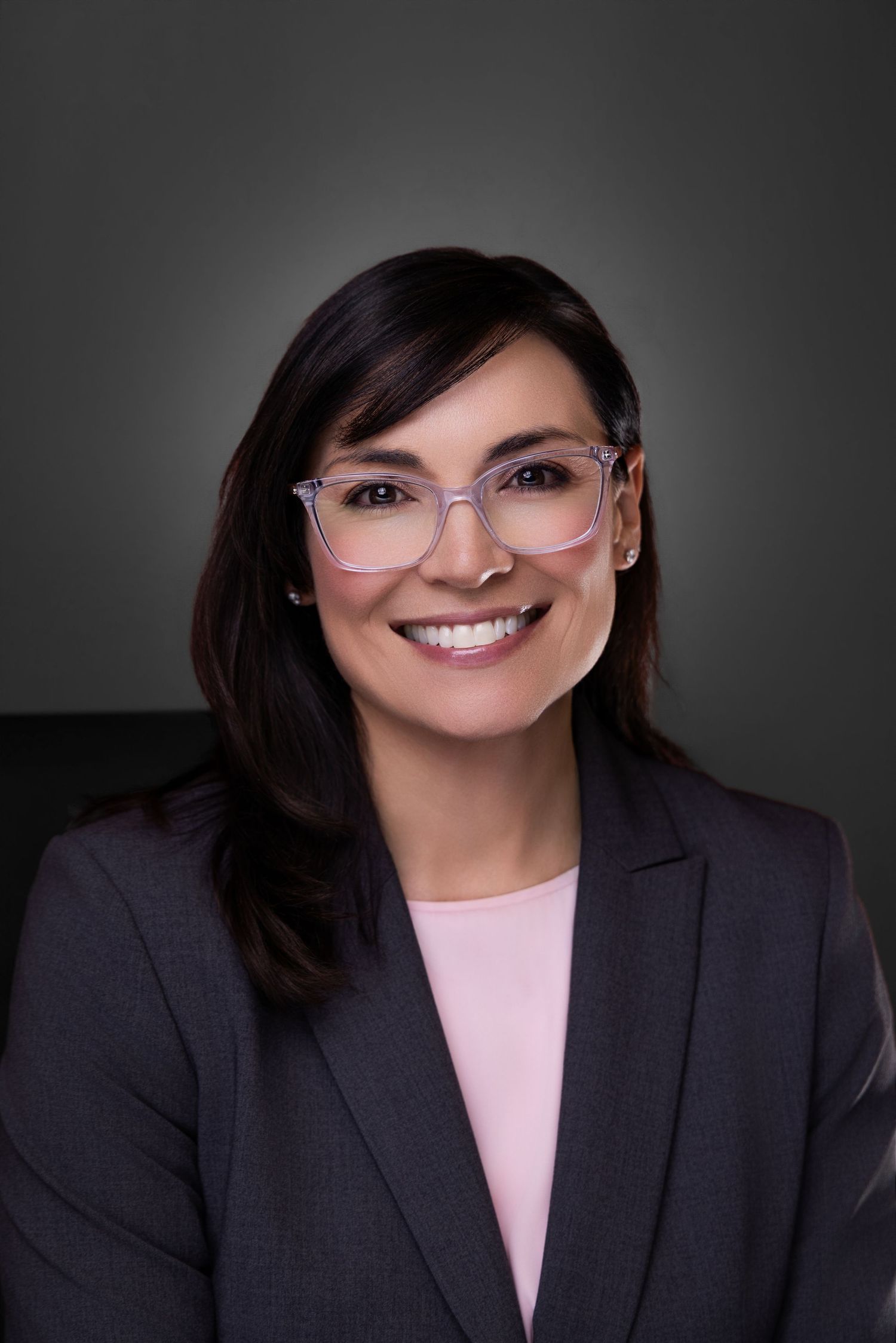 Professional headshot of smiling brunette businesswoman wearing clear-framed glasses and gray blazer on dark background.