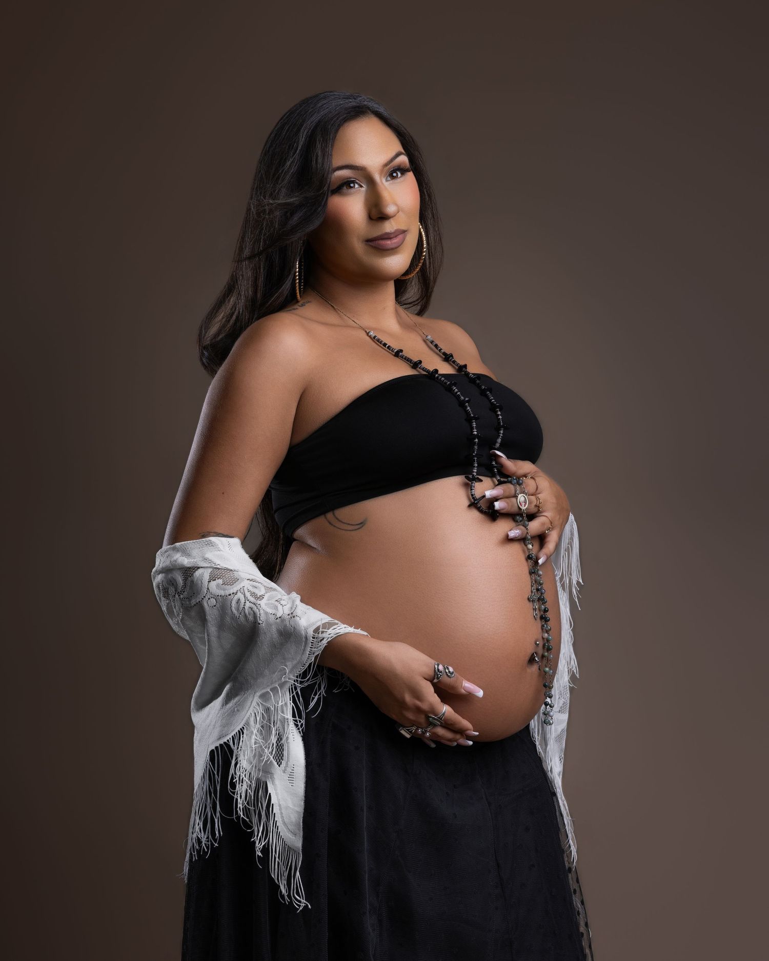Pregnant woman poses in black bandeau top with rosary beads showcasing baby bump in studio.