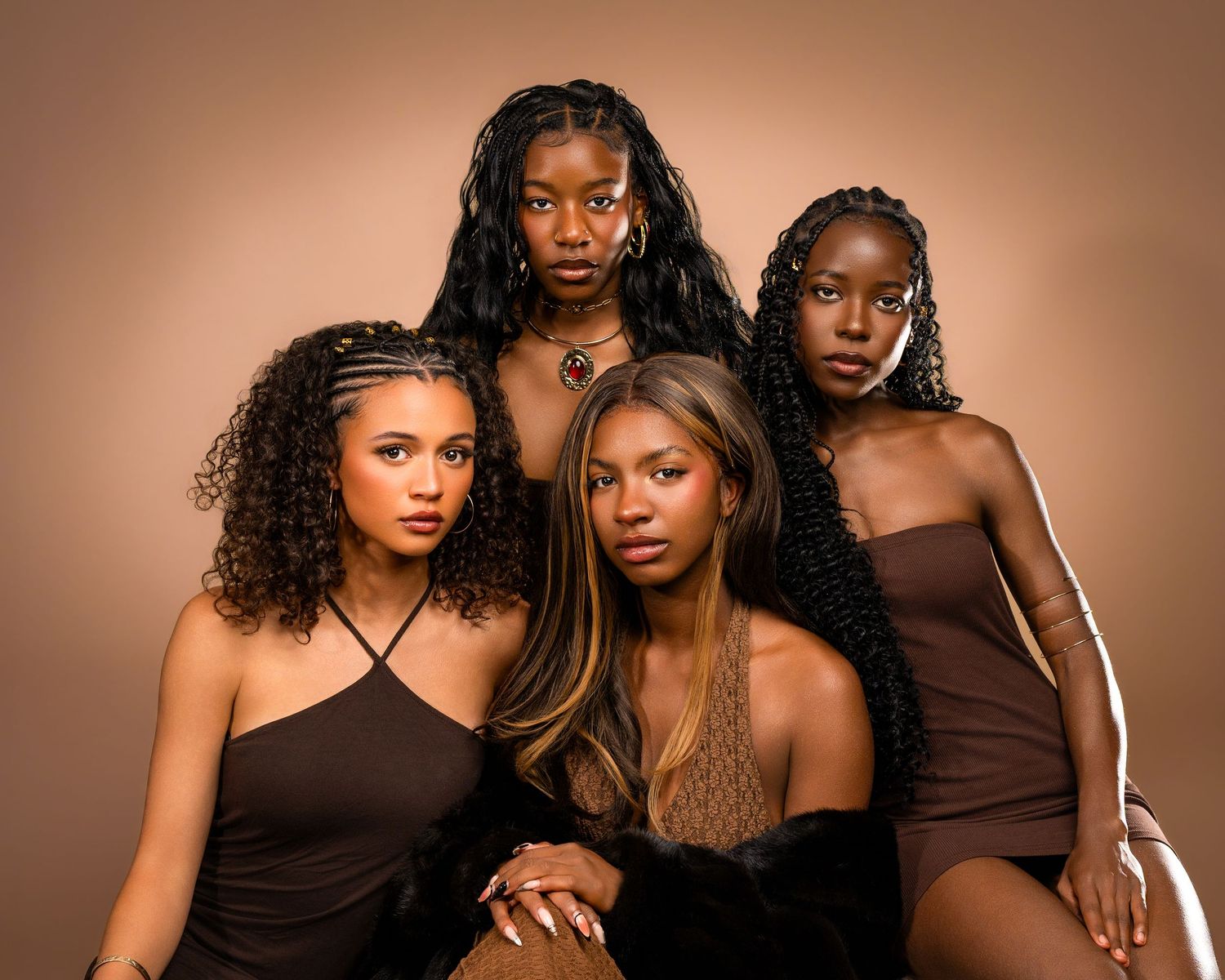 Four young women in brown outfits pose together against a warm tan background in a professional photo shoot.