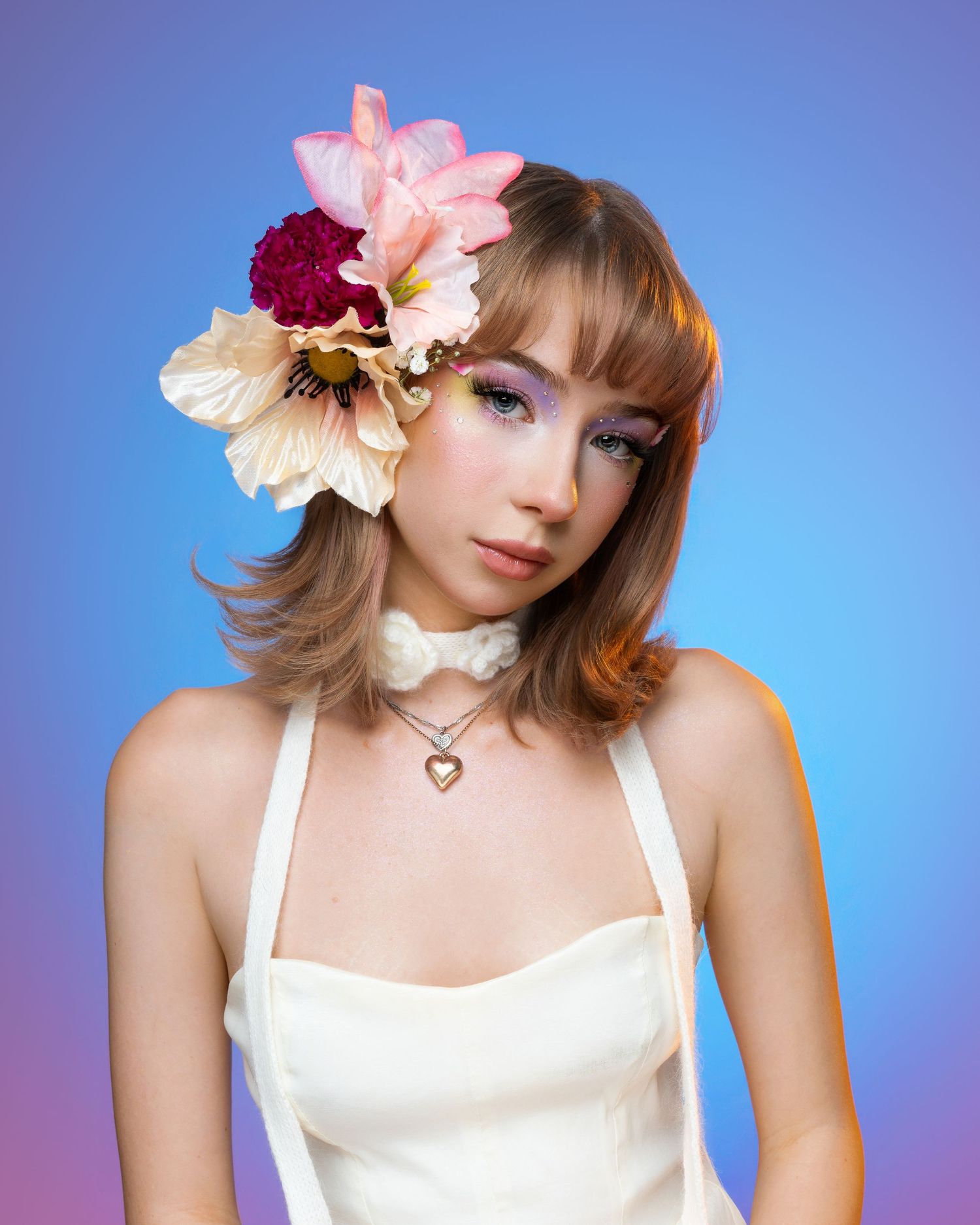 Young woman in white dress with floral hair accessories and heart necklace poses against blue background.