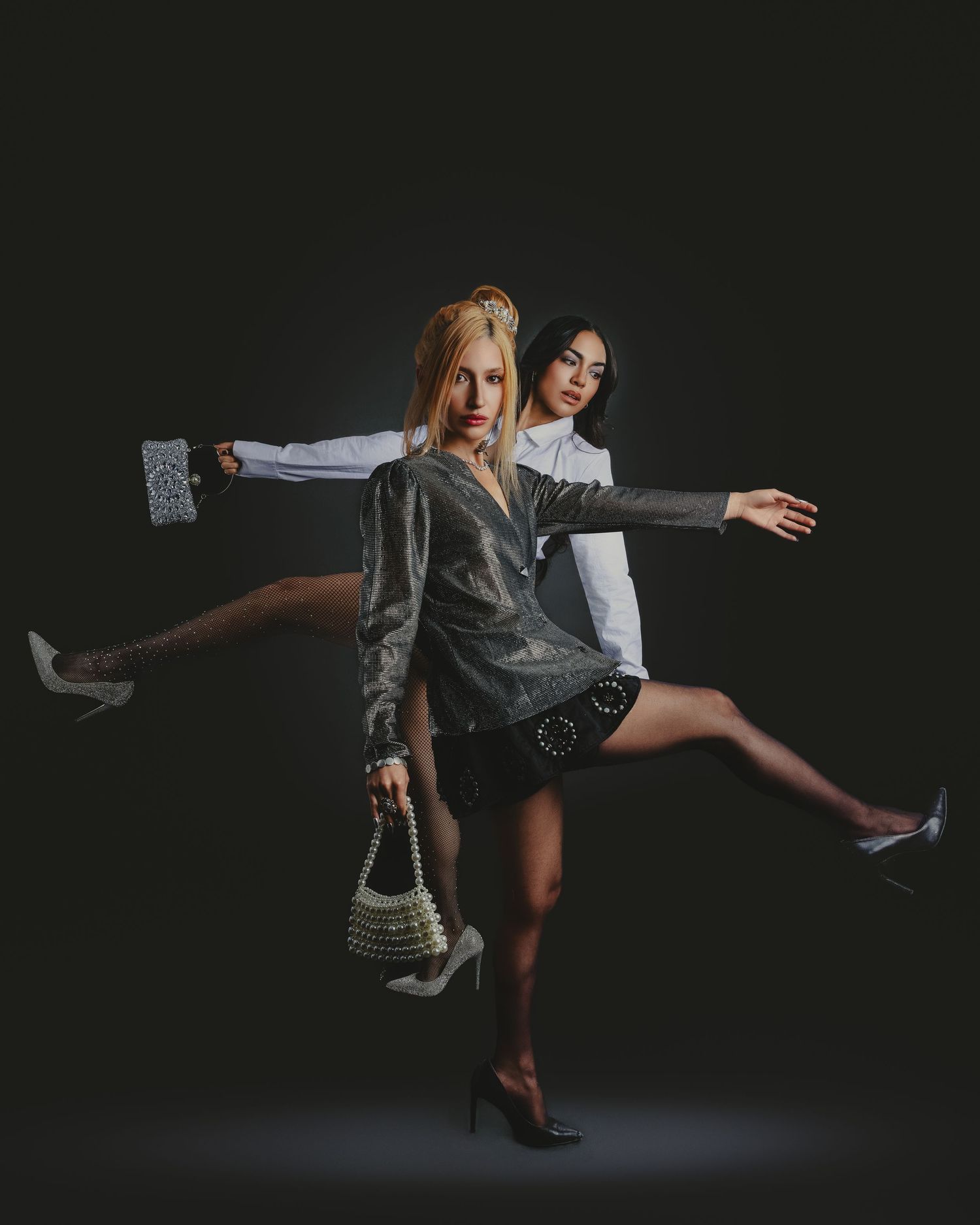 Two stylish women pose dynamically in elegant outfits, one in silver velvet, against a dark studio background.