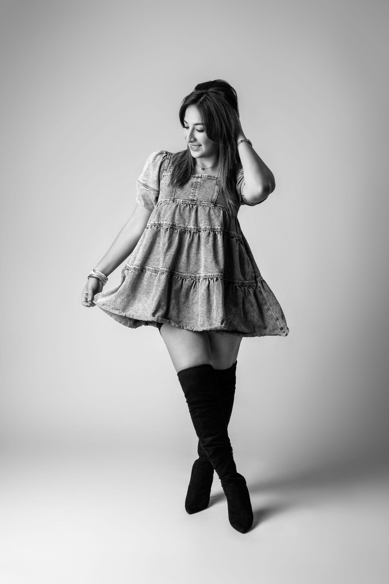 Black and white photo of a young woman in a short tiered dress and thigh-high boots, posing gracefully in a studio.