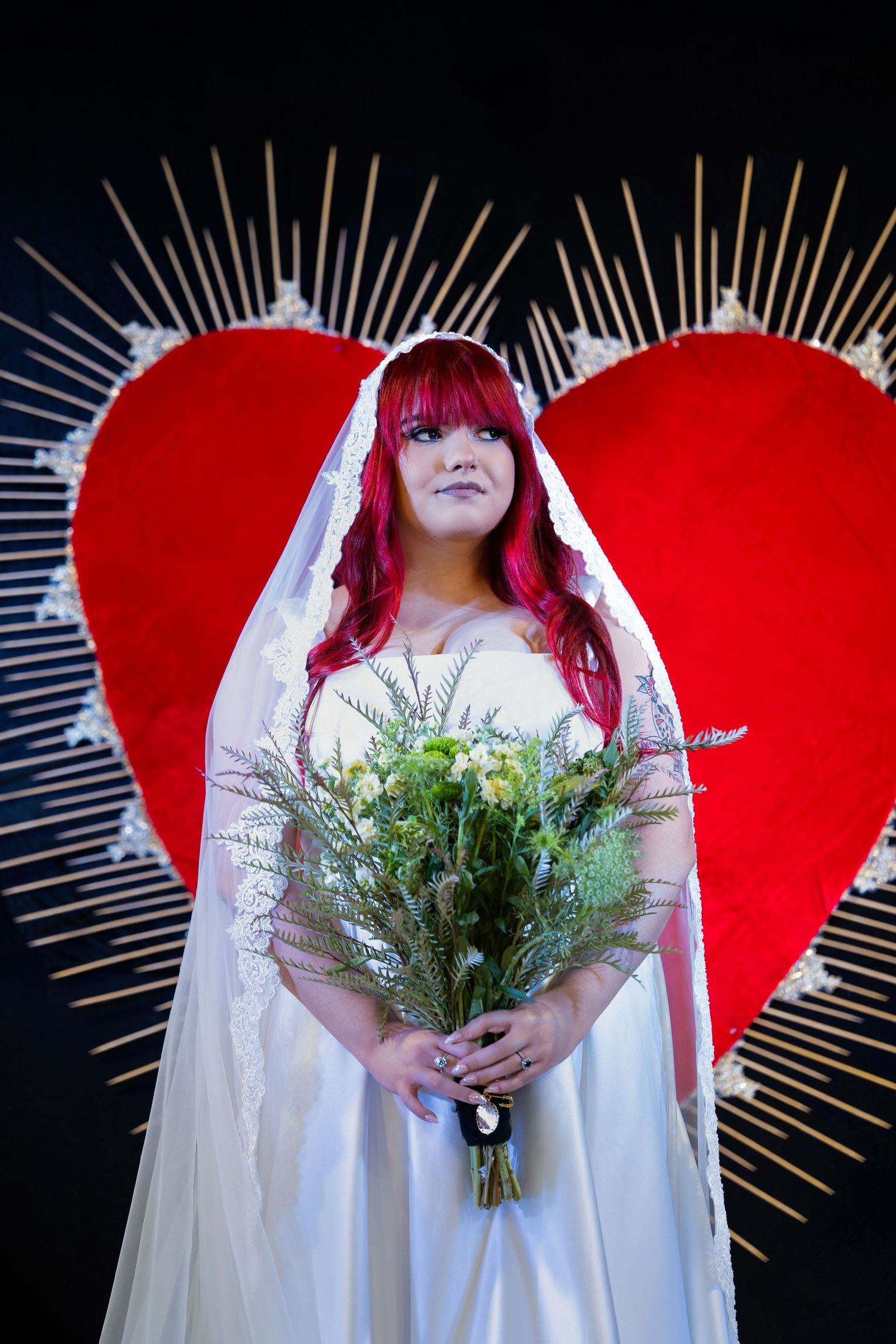 Red-haired bride in white gown holds wildflower bouquet before large red heart backdrop with golden rays.