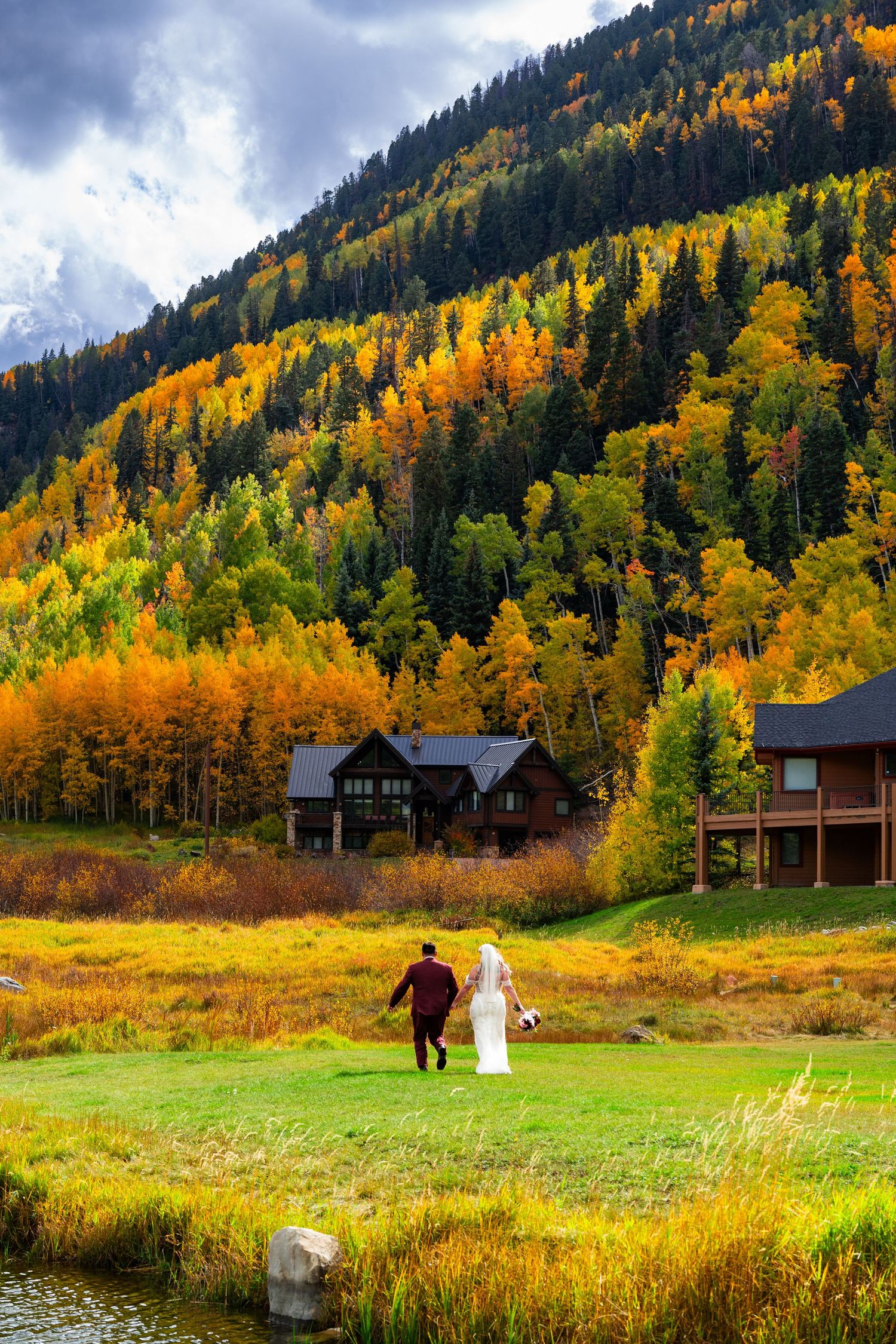 Newlyweds walk hand-in-hand across green meadow with stunning autumn mountain foliage backdrop.