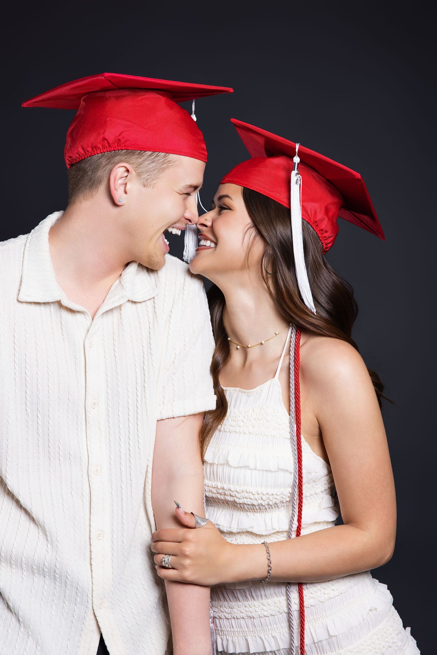 Laughing couple in red graduation caps sharing a joyful moment together in a dark studio.