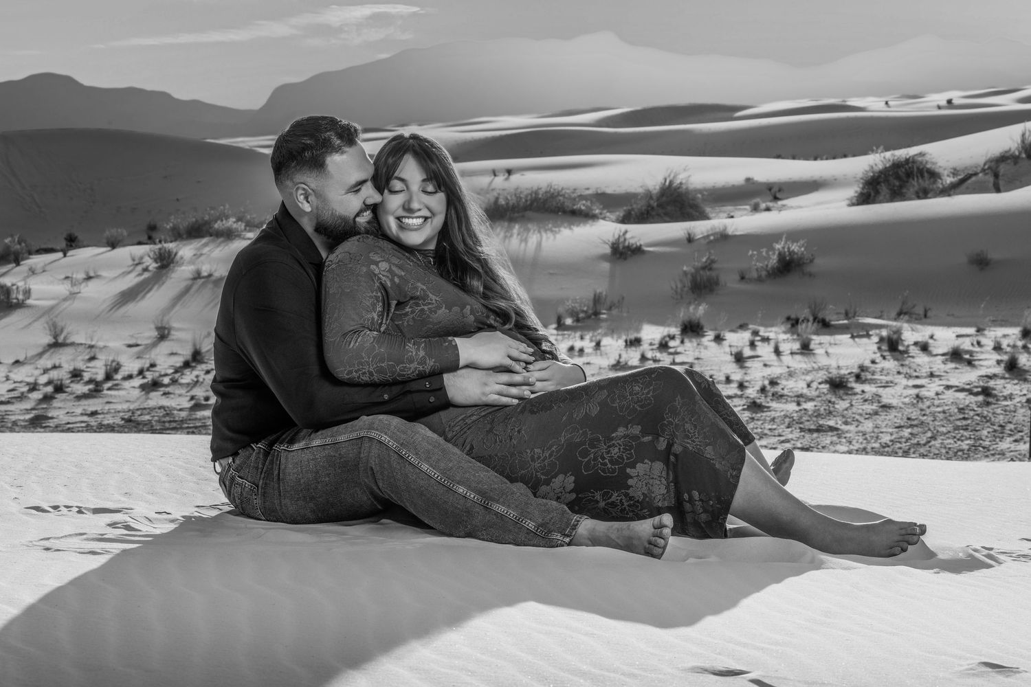 Black and white photo of expecting couple sitting together on white sand dunes with scenic desert landscape behind them.
