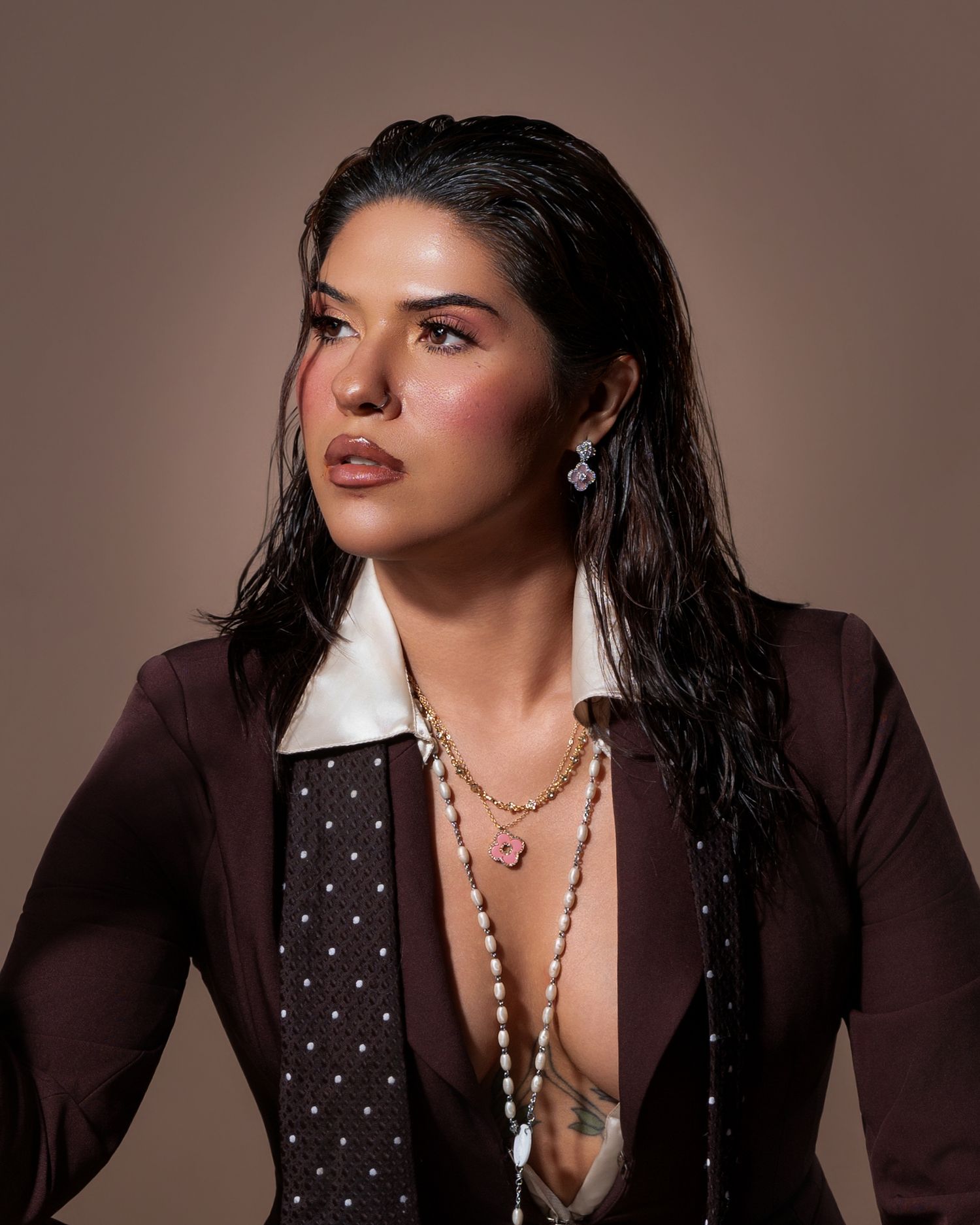 Woman in brown blazer with white collar, layered necklaces, and pink earrings poses against a warm beige background.