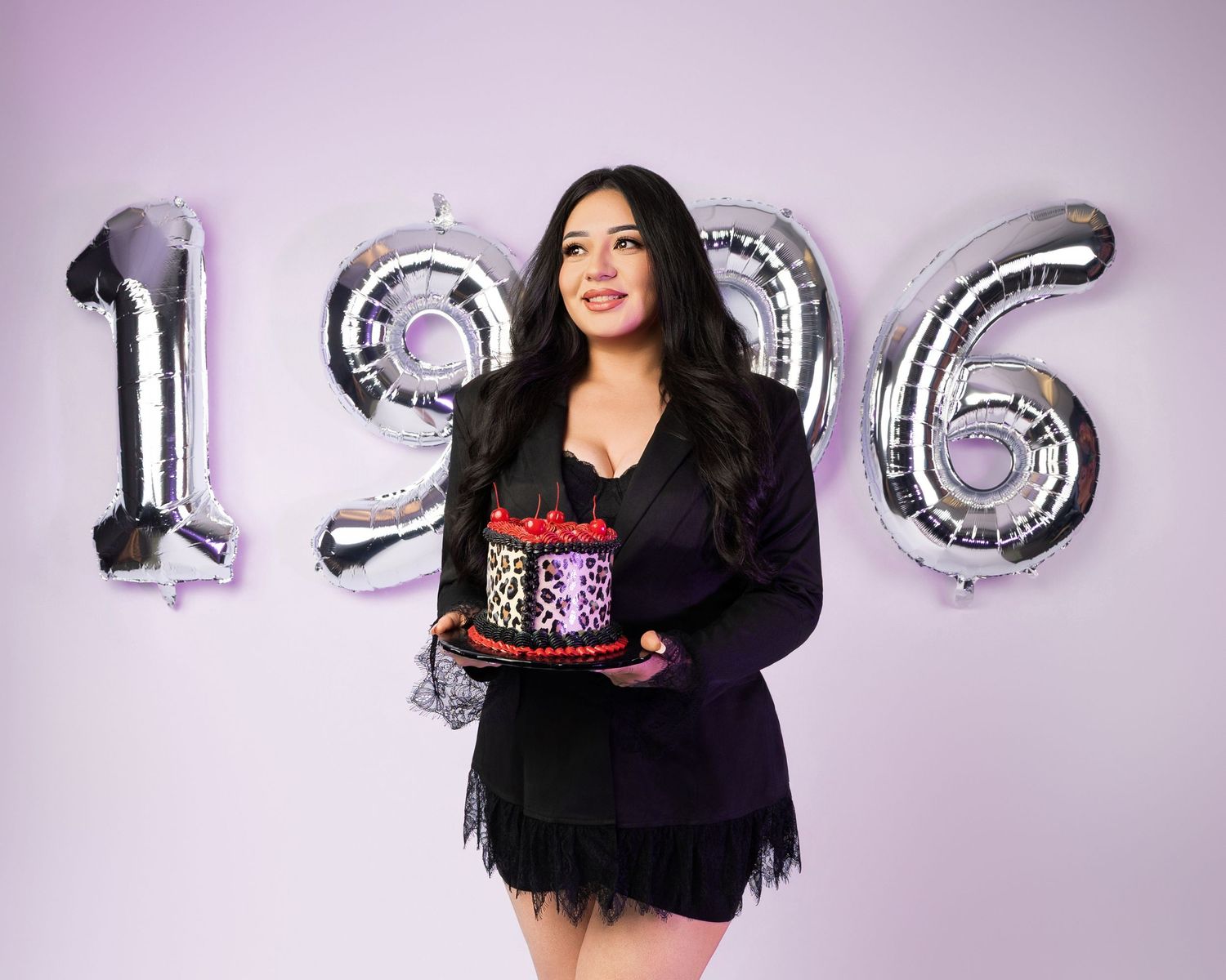 Woman in black outfit holding decorated cake, posing with silver balloon numbers 1996 against purple background.