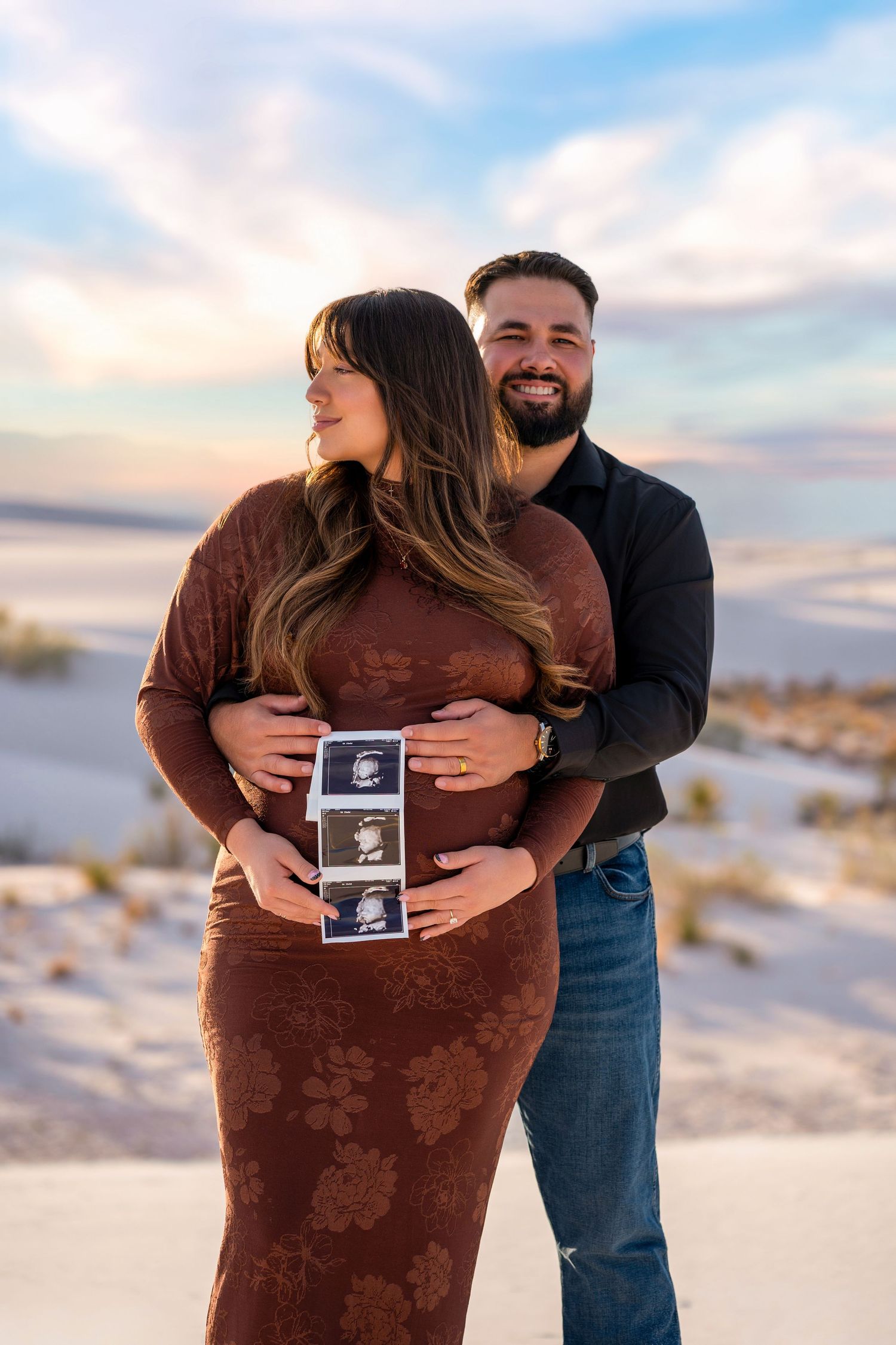 Couple holding ultrasound photos at White Sands, pregnant woman in brown floral dress at sunset.