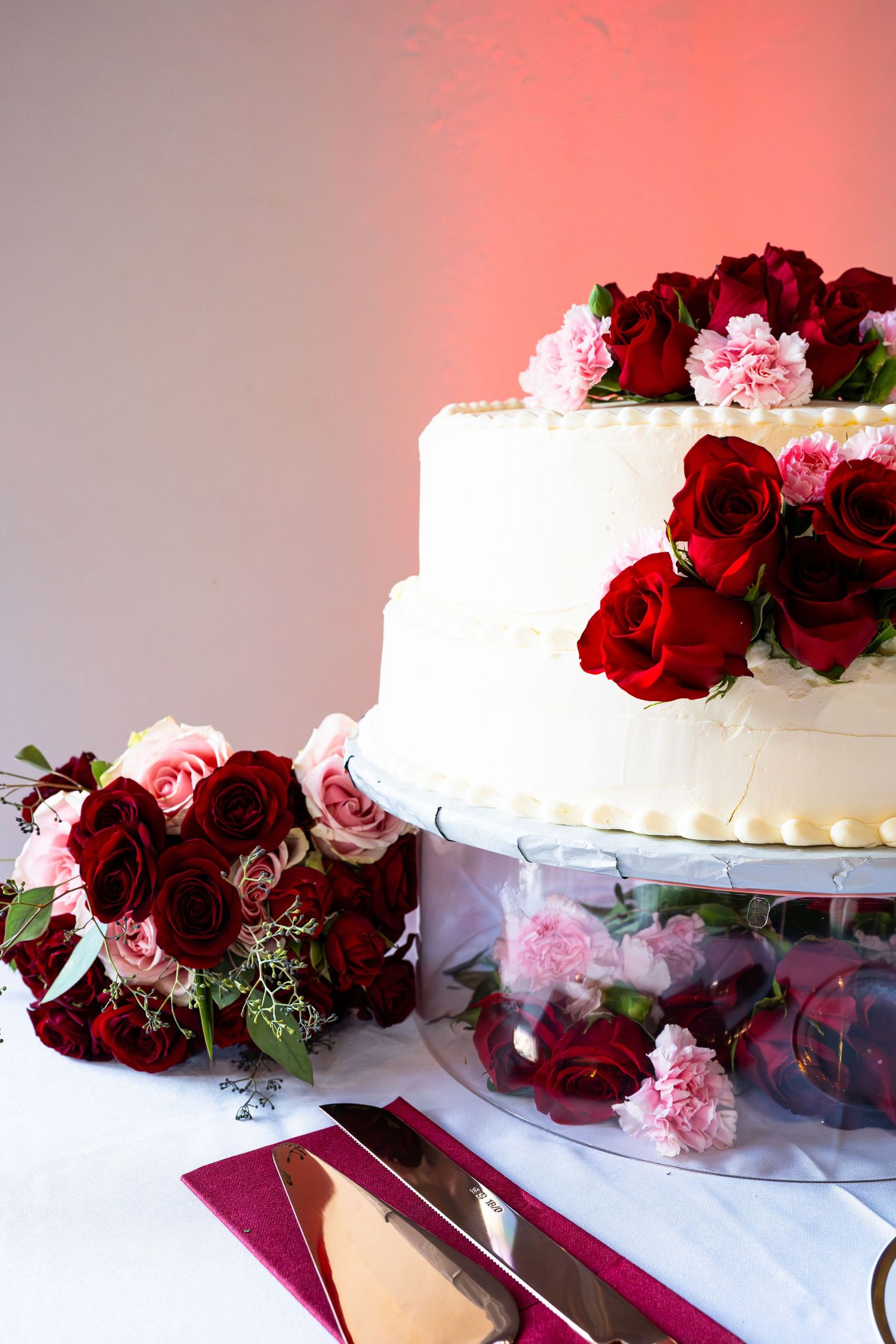 Two-tier white wedding cake decorated with red roses and pink carnations on reception table.