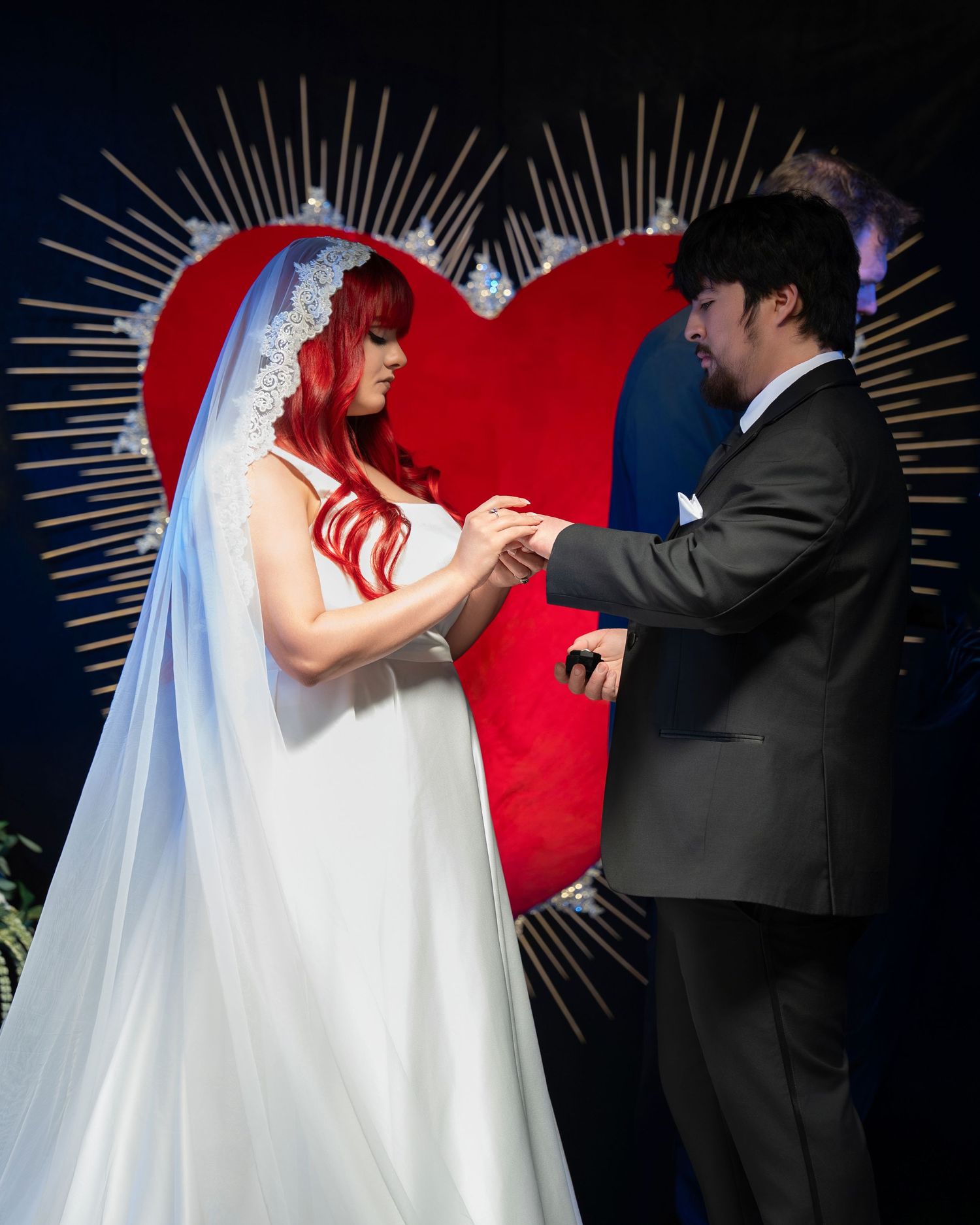 Red-haired bride and groom exchange rings before a large red heart backdrop during wedding ceremony.
