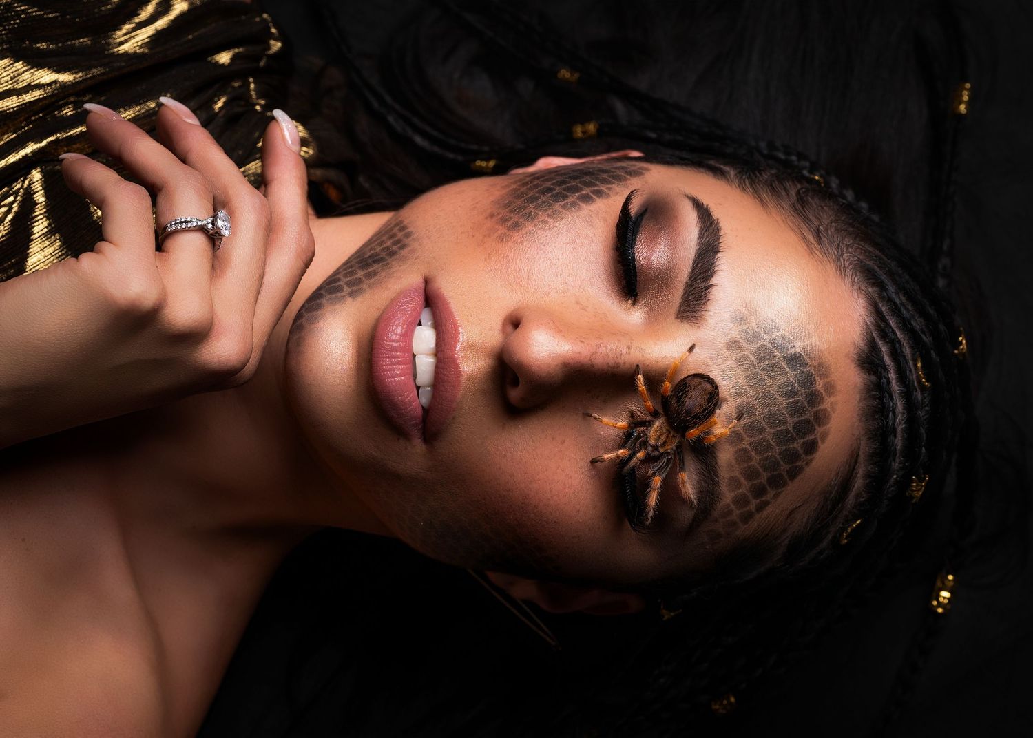 A woman with braids and dramatic makeup poses with a spider on a veil over her face against a dark background.