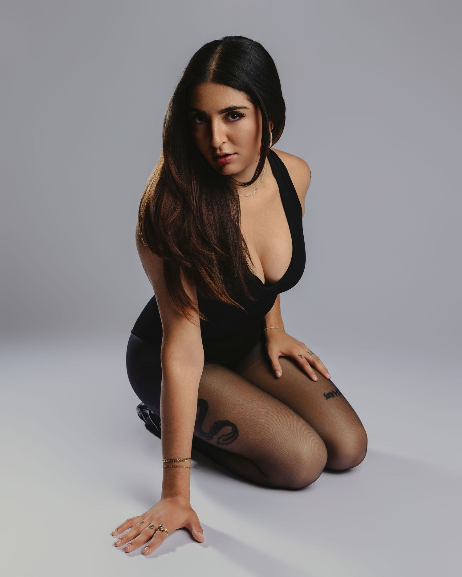 Woman in black bodysuit and sheer tights poses seated on floor with long dark hair and gold accessories.