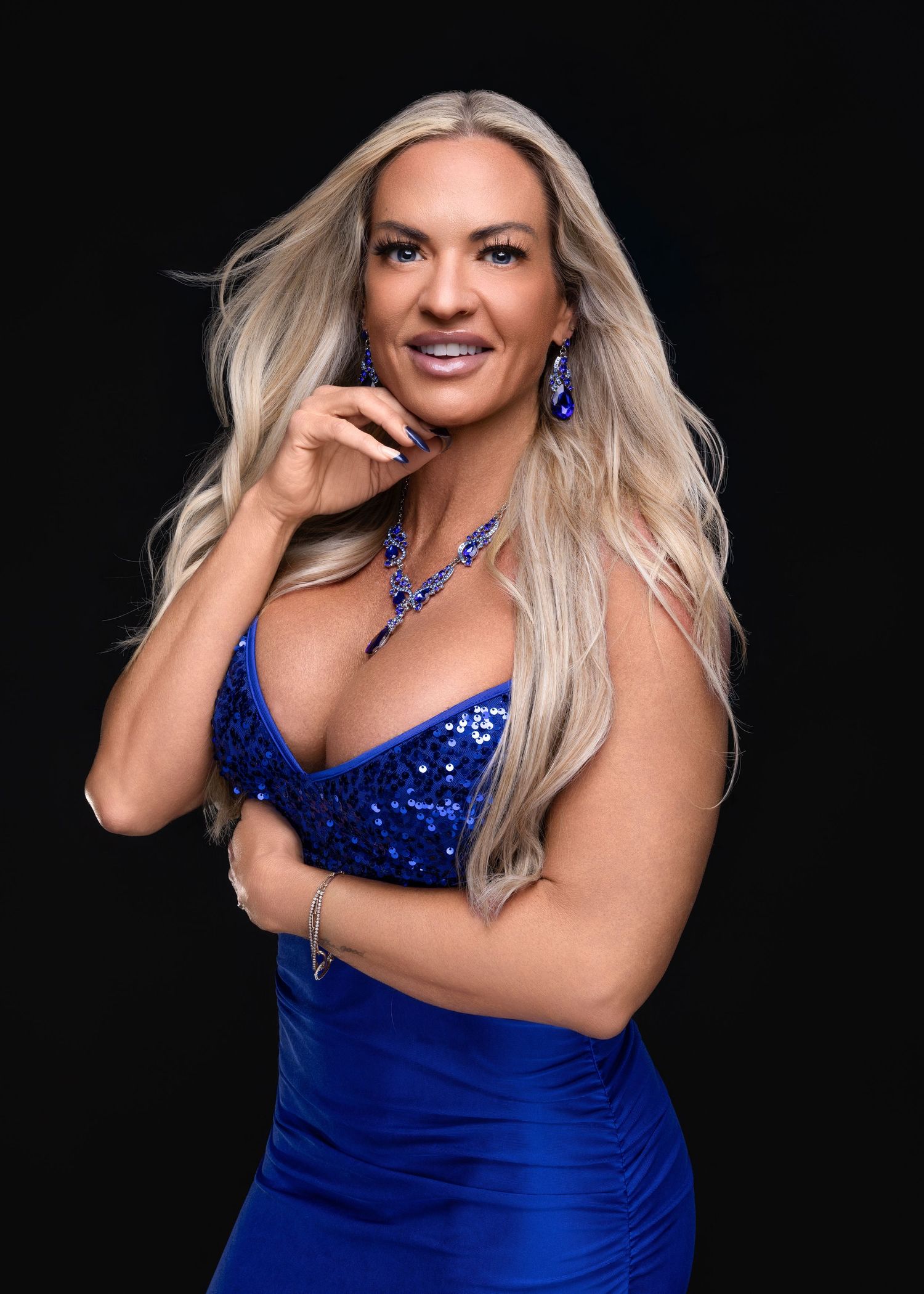 Blonde woman in a royal blue sequin dress with matching sapphire jewelry poses confidently against a dark background.